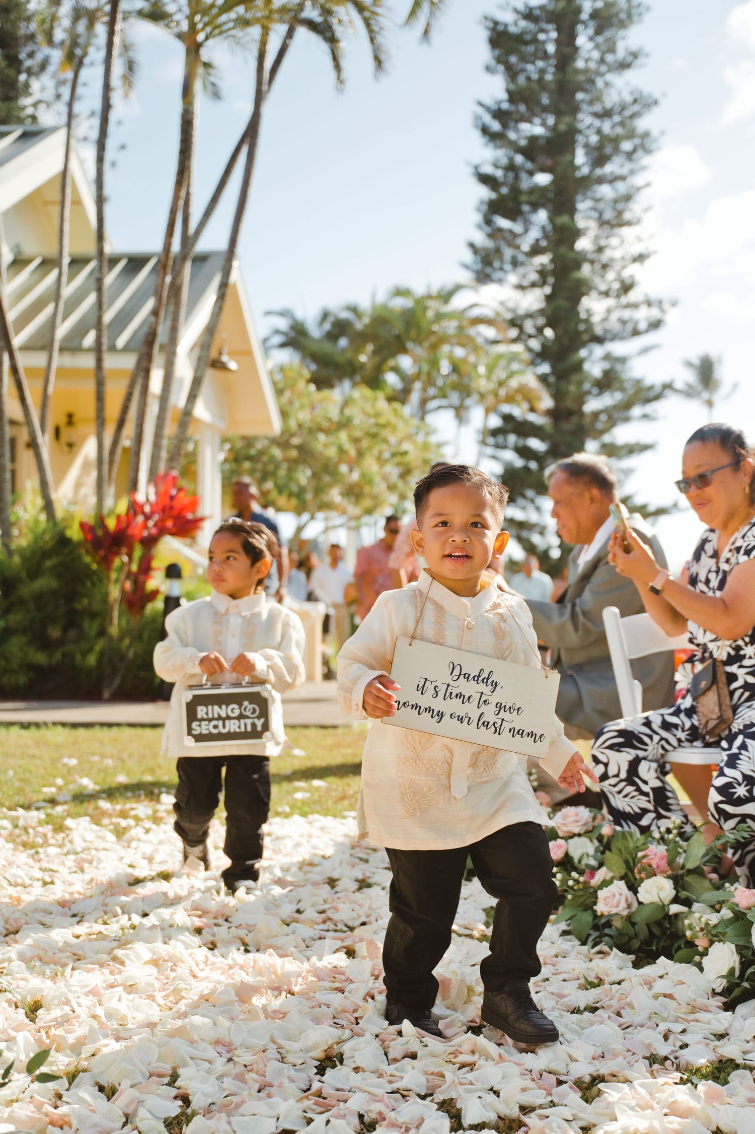 Two young ring bearers walk down a flower petal aisle at an outdoor wedding, holding signs for the ceremony.