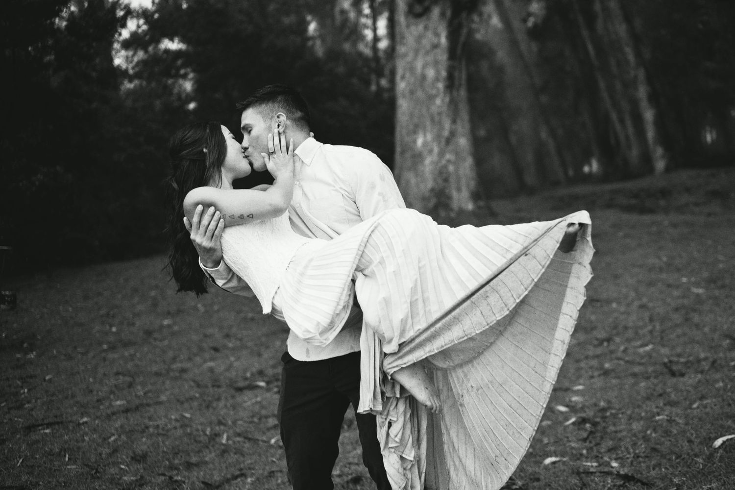 Groom lifting bride in white dress sharing a kiss in a black and white forest wedding photo.