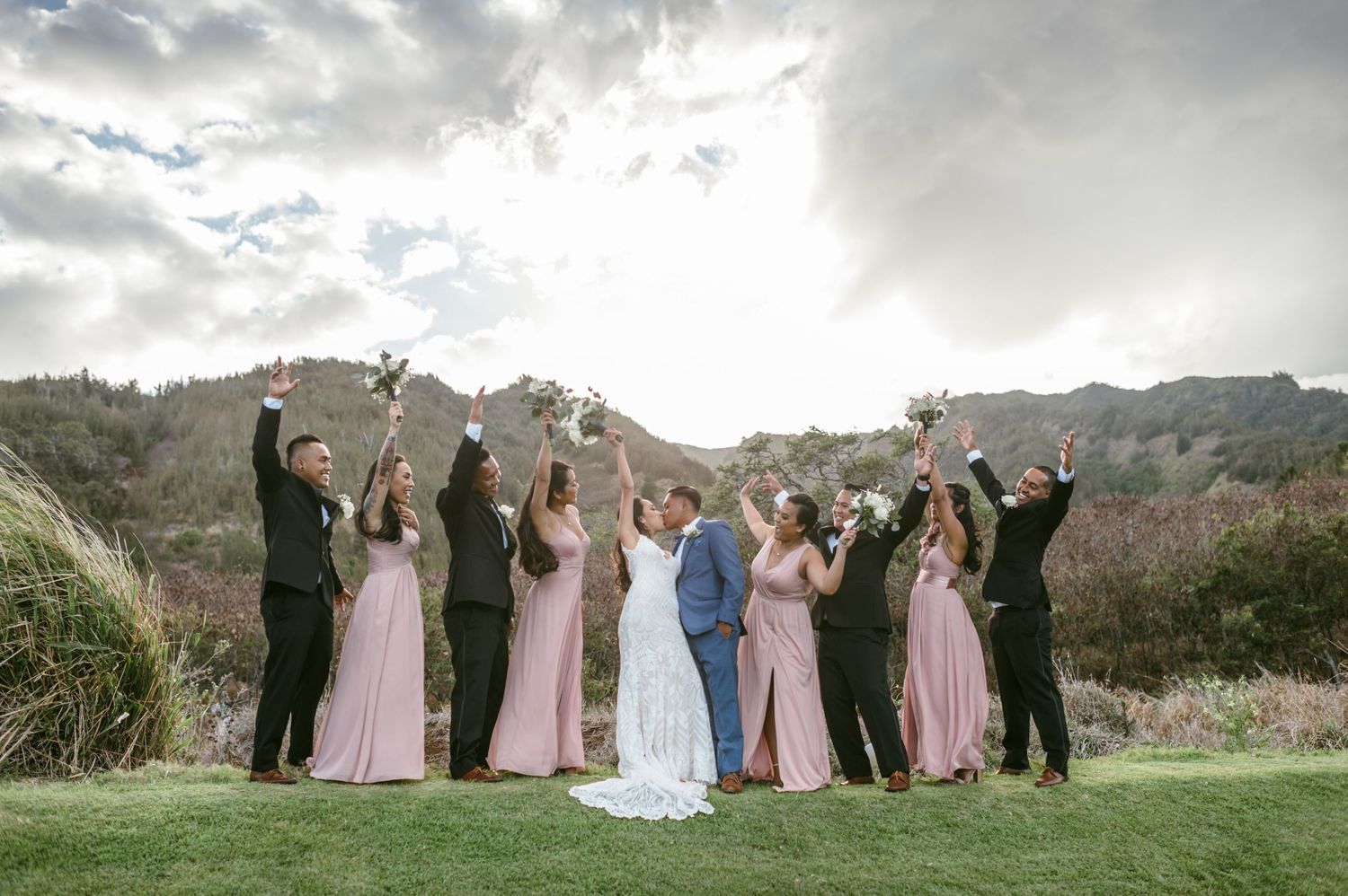 Wedding party celebrates with arms raised against mountainous backdrop at outdoor venue.