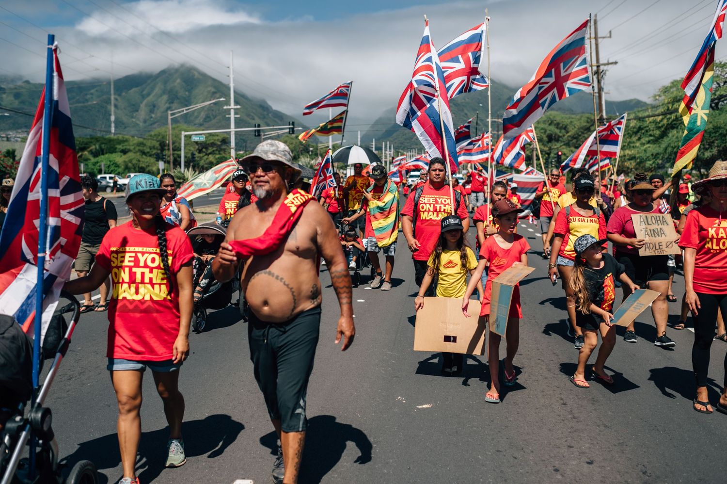 Demonstrators march with Hawaiian state flags along a road with green mountains visible in the distance.