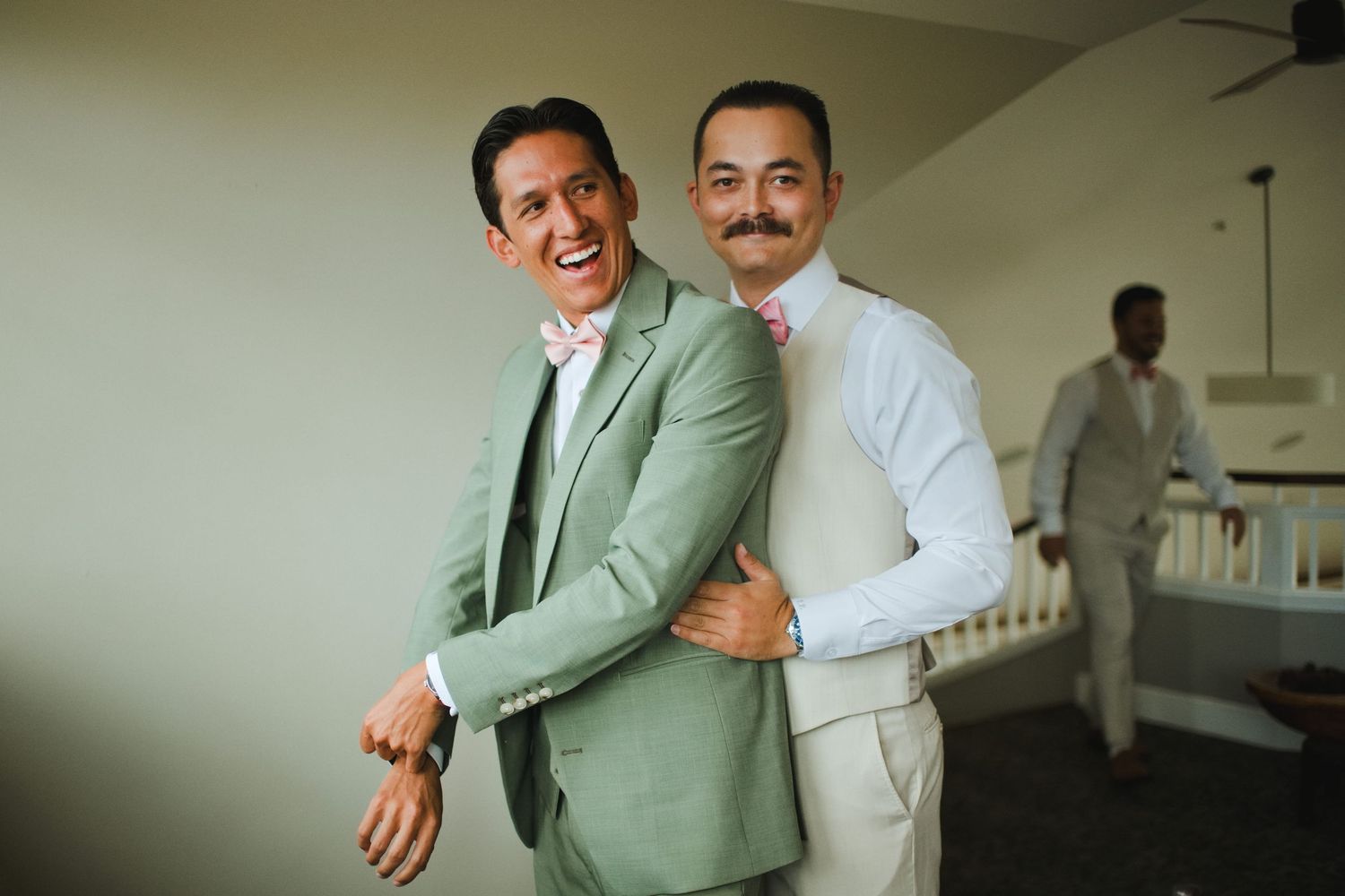 Two men in elegant wedding attire, one in sage green suit, share a joyful embrace while smiling at the camera.