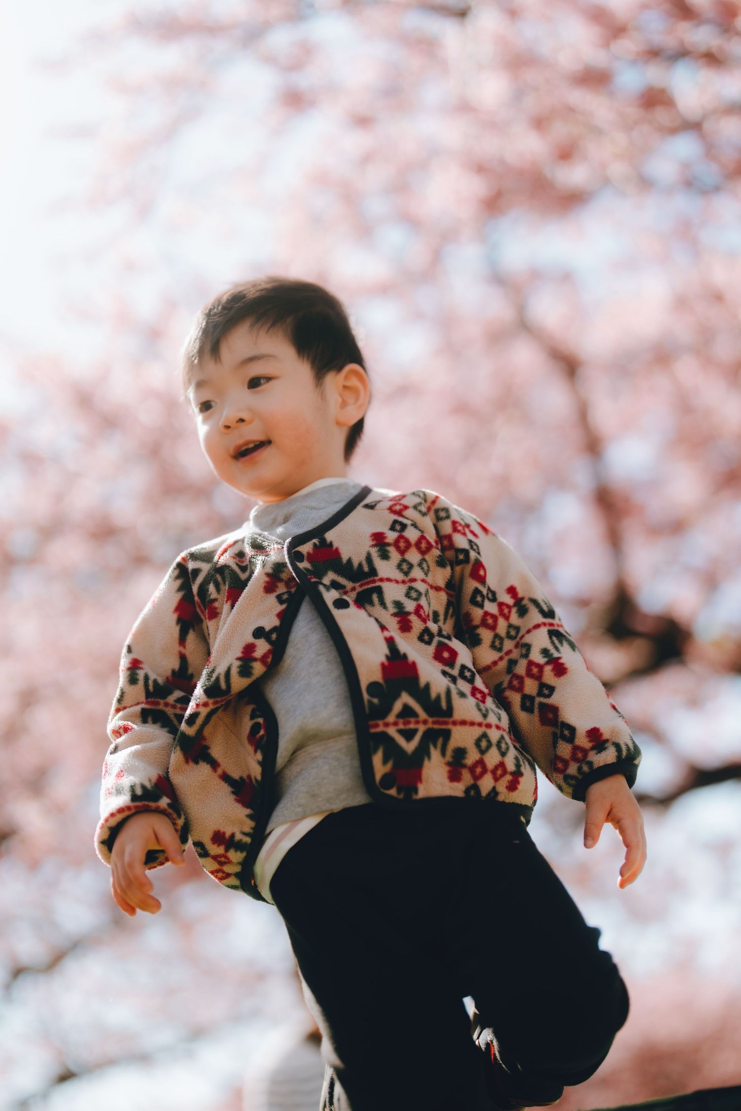 A young Asian boy in a colorful patterned jacket smiles outdoors under blooming cherry blossom trees in spring.