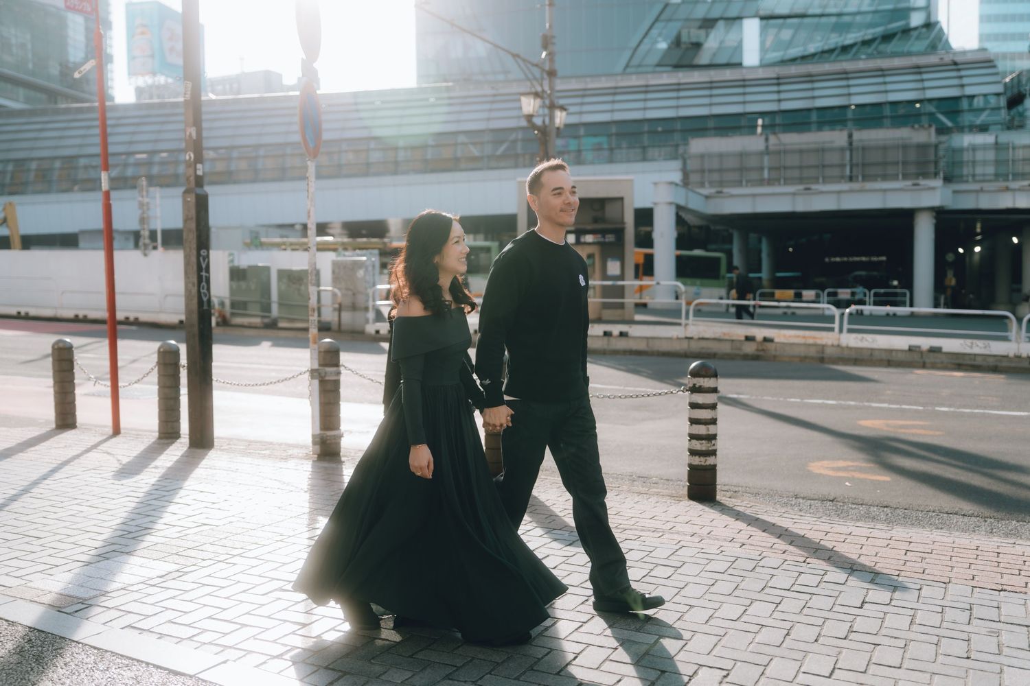Couple walking together in matching black outfits on a sunny shibuya street with modern buildings.
