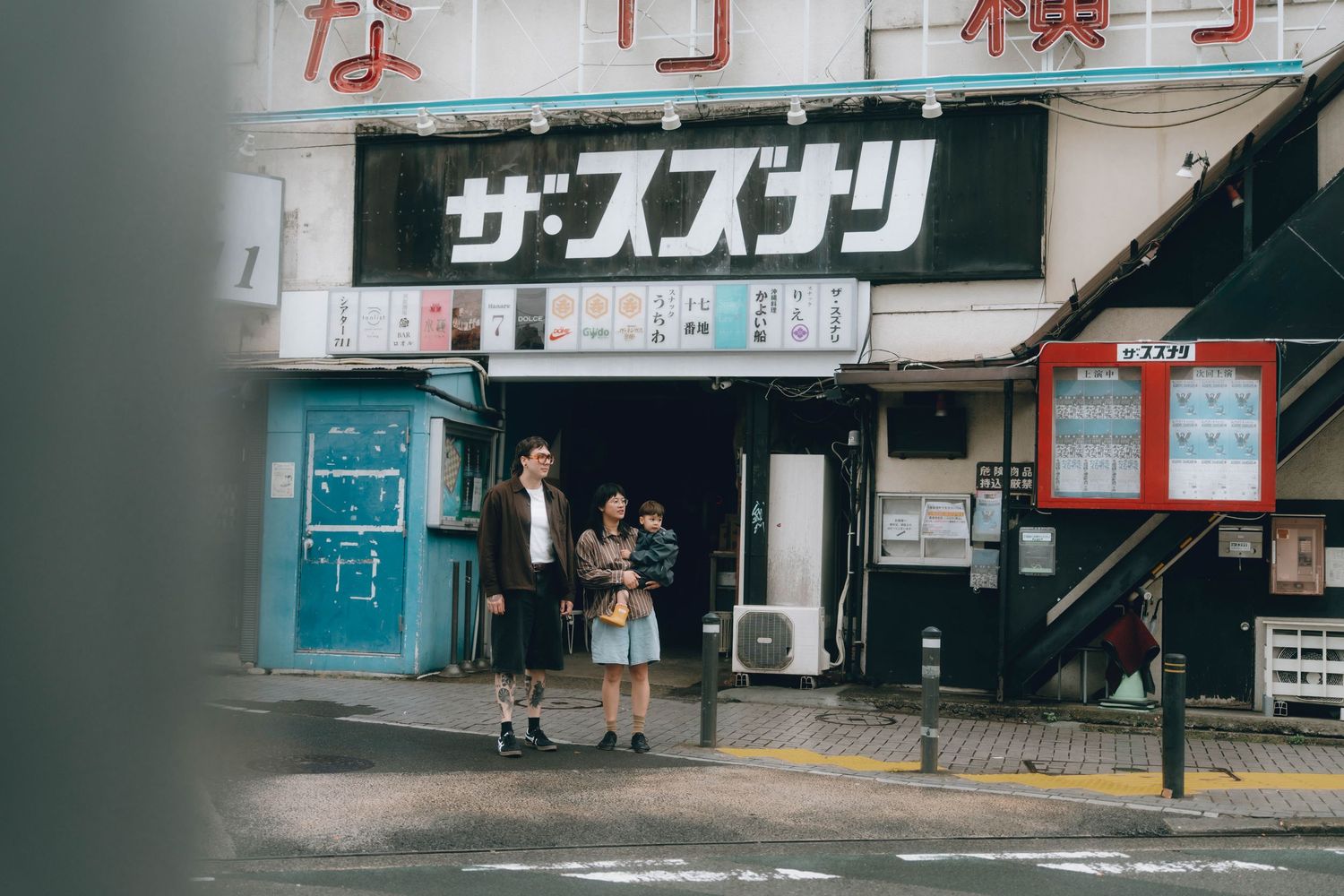 Two people walking outside a Japanese theater building with kanji signage in an urban street scene in Shimokitazawa, Tokyo.