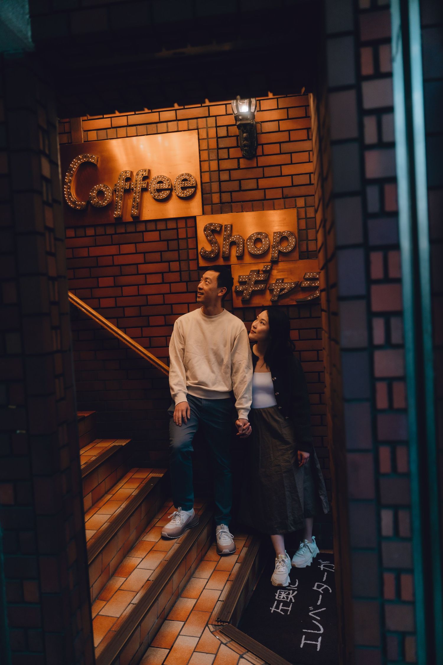 Couple holding hands outside a cozy coffee shop entrance with warm brick walls and Japanese signage at night.