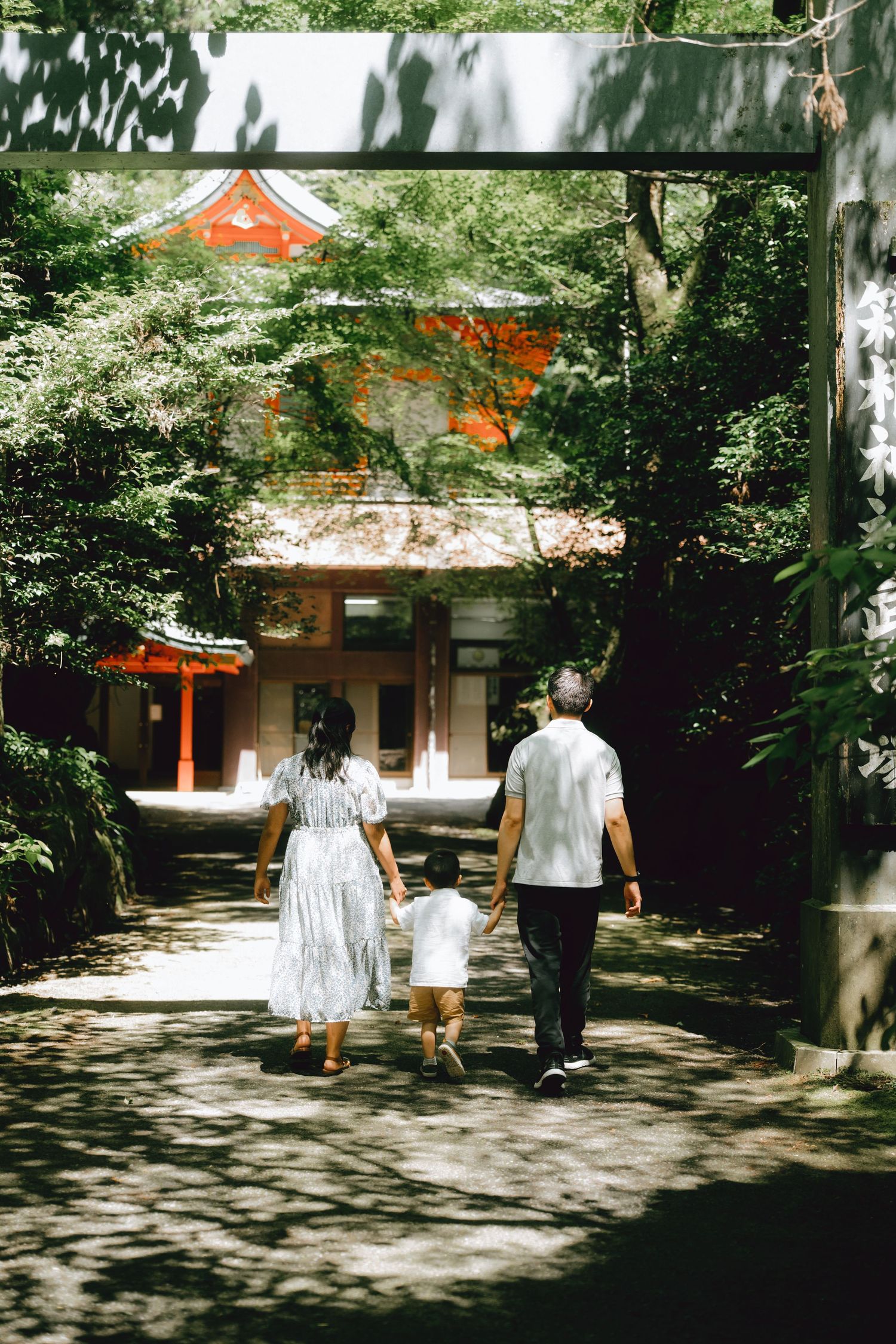 Family walking hand-in-hand along a shaded path toward a traditional red Japanese shrine surrounded by lush green trees.