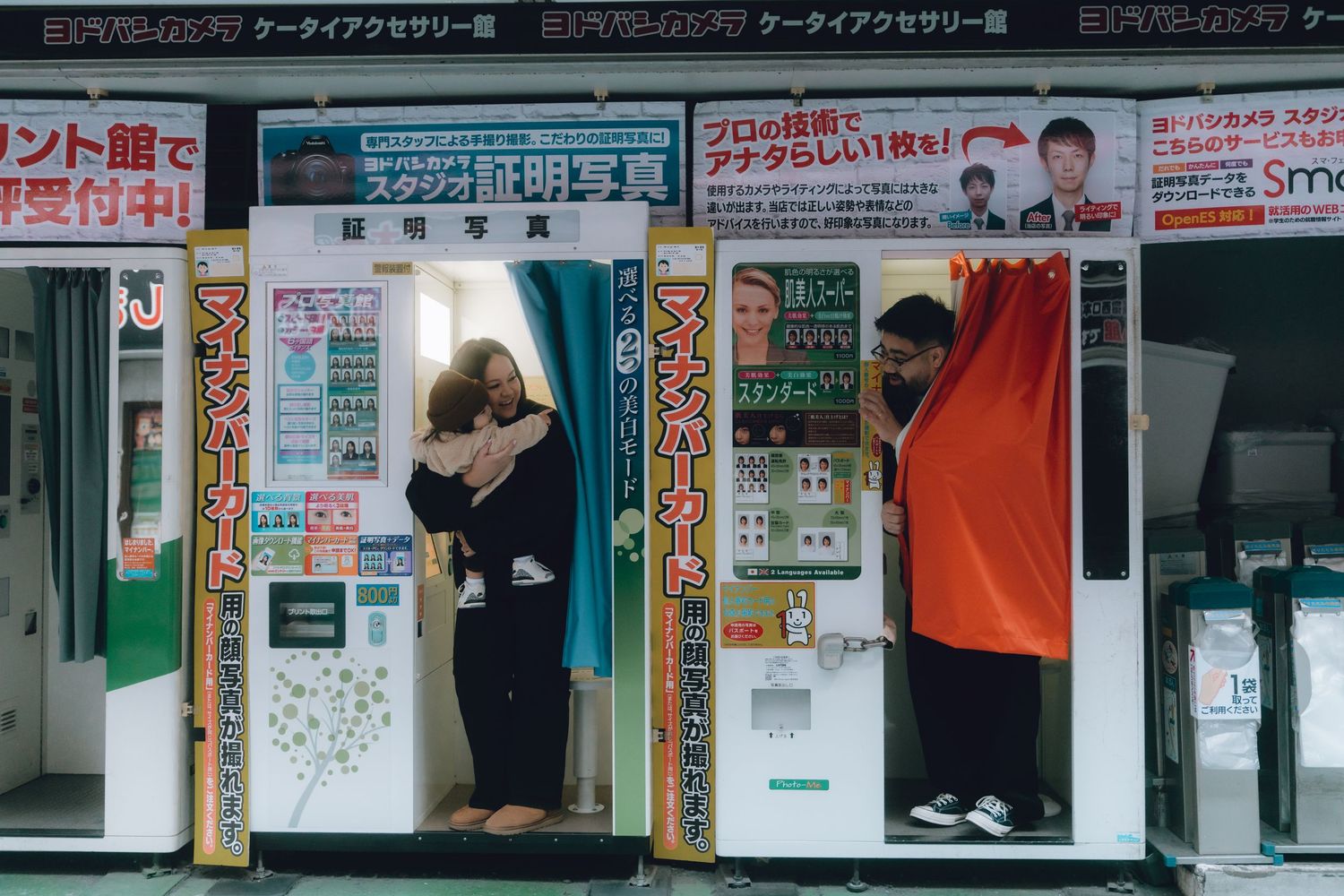 Two people using a photo booth machine on a Japanese street, with colorful signage and a red curtain visible.