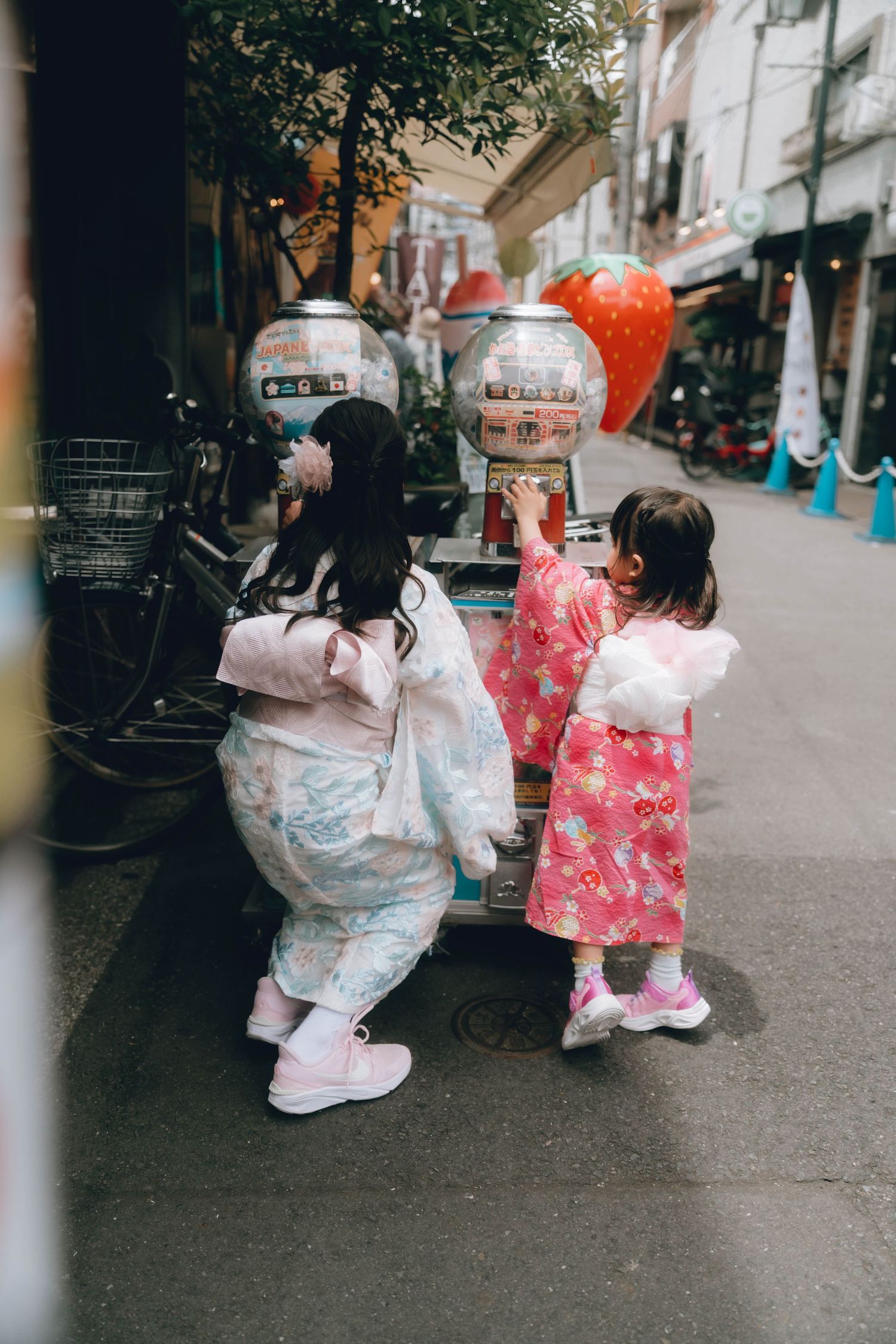 Two young girls in colorful kimonos use a gumball machine on a charming Japanese street in Aasakusa.
