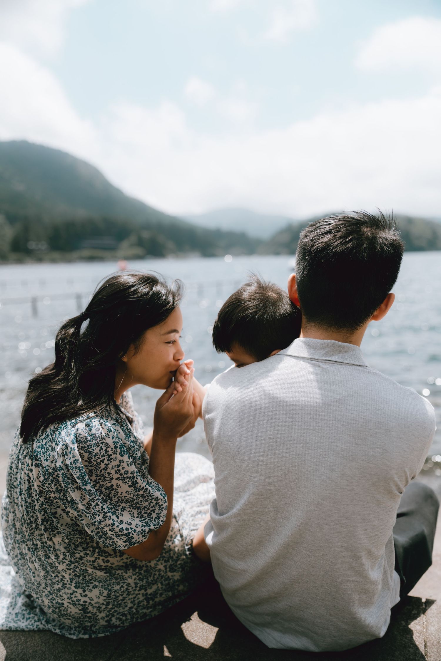 Family of three sitting by a mountain lake, mother kissing child's hand while father holds the toddler outdoors in Lake Ashi Hakone,
