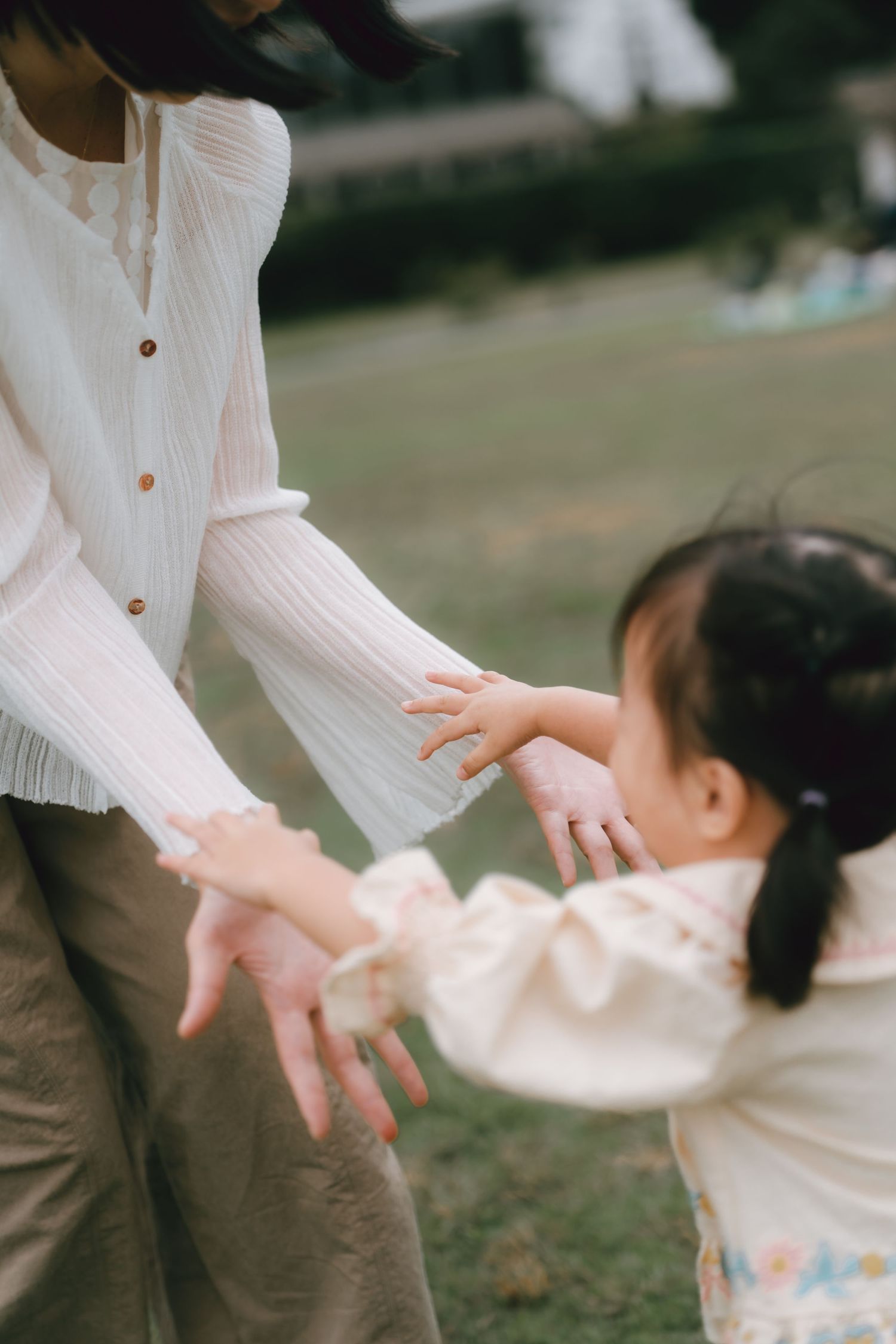 A young Asian girl reaches out to hold hands with her mother in a sunlit park, both wearing soft white outfits.