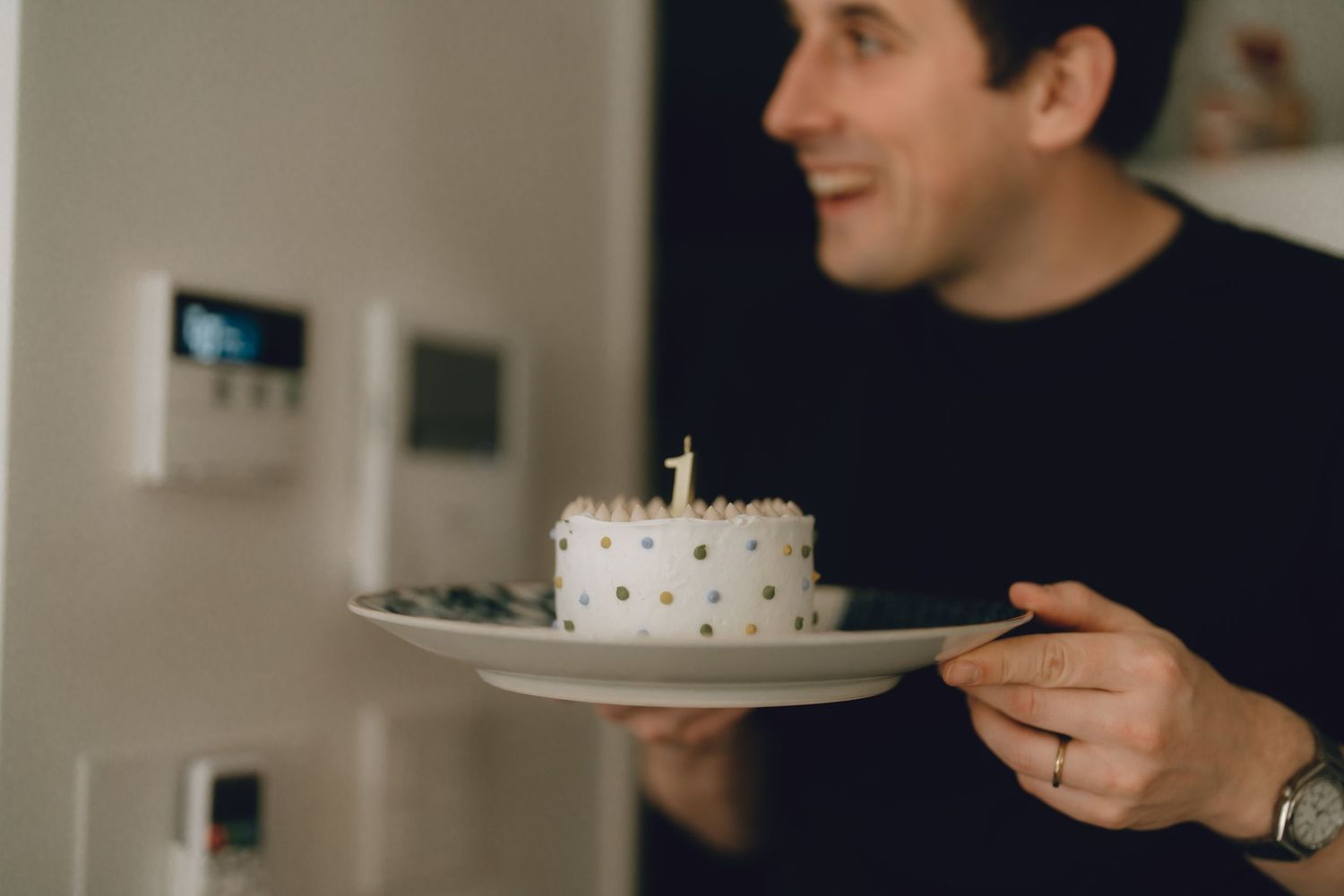 Man smiling while holding a white birthday cake with number 1 candle on a plate indoors.