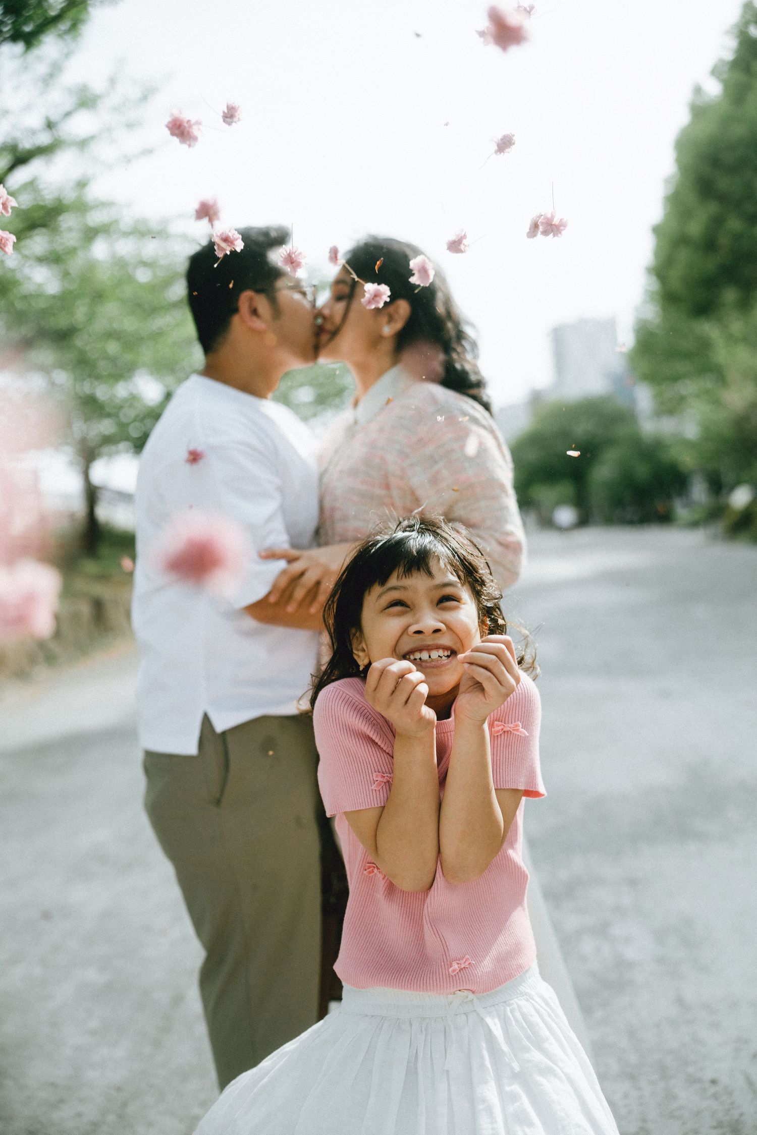 A young girl smiles joyfully as cherry blossom petals fall around her while her parents share a kiss behind her in asakusa.