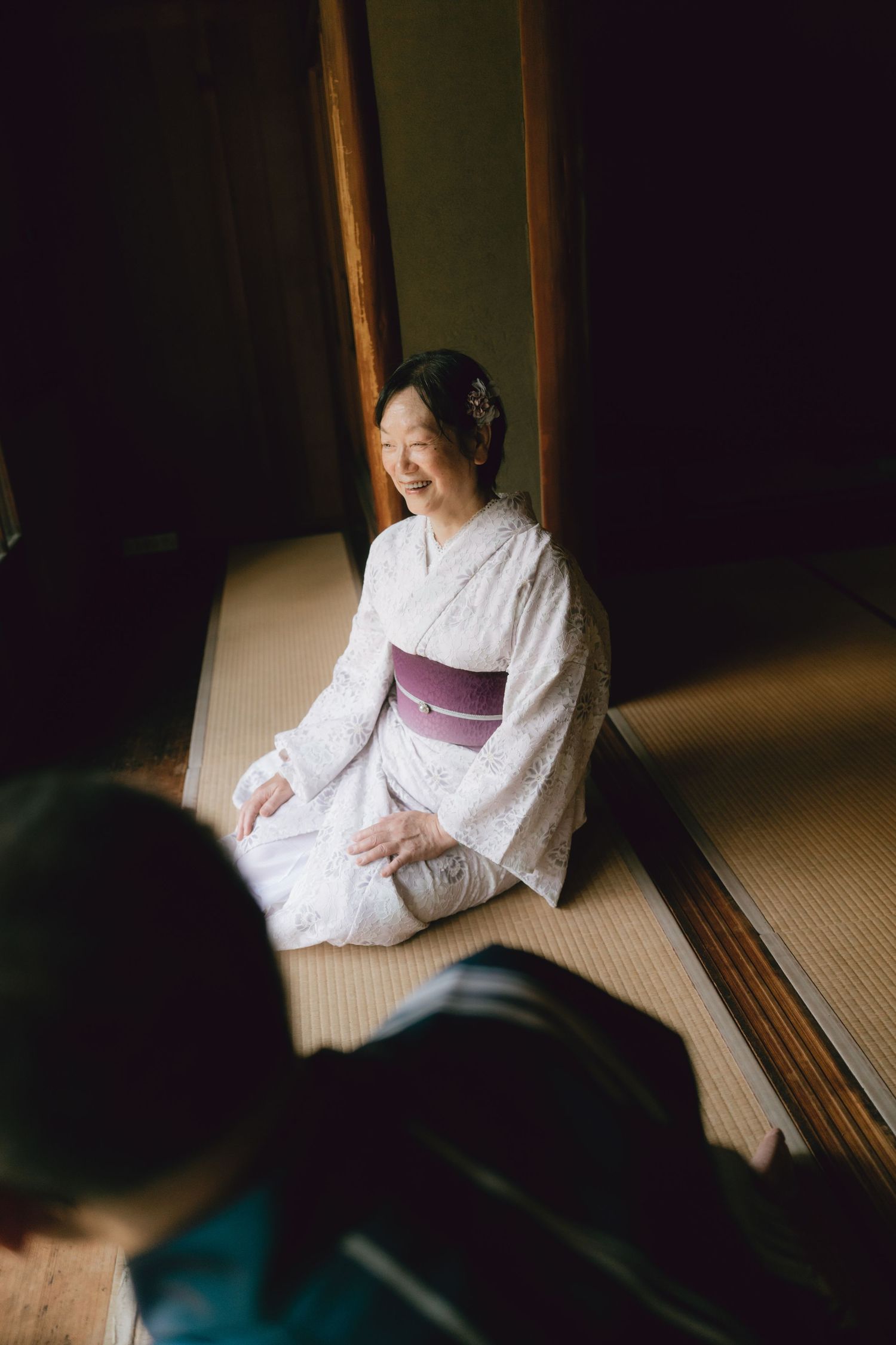 A smiling woman in a white kimono with purple obi sits gracefully on tatami mats in a traditional Japanese room. in kawagoe.