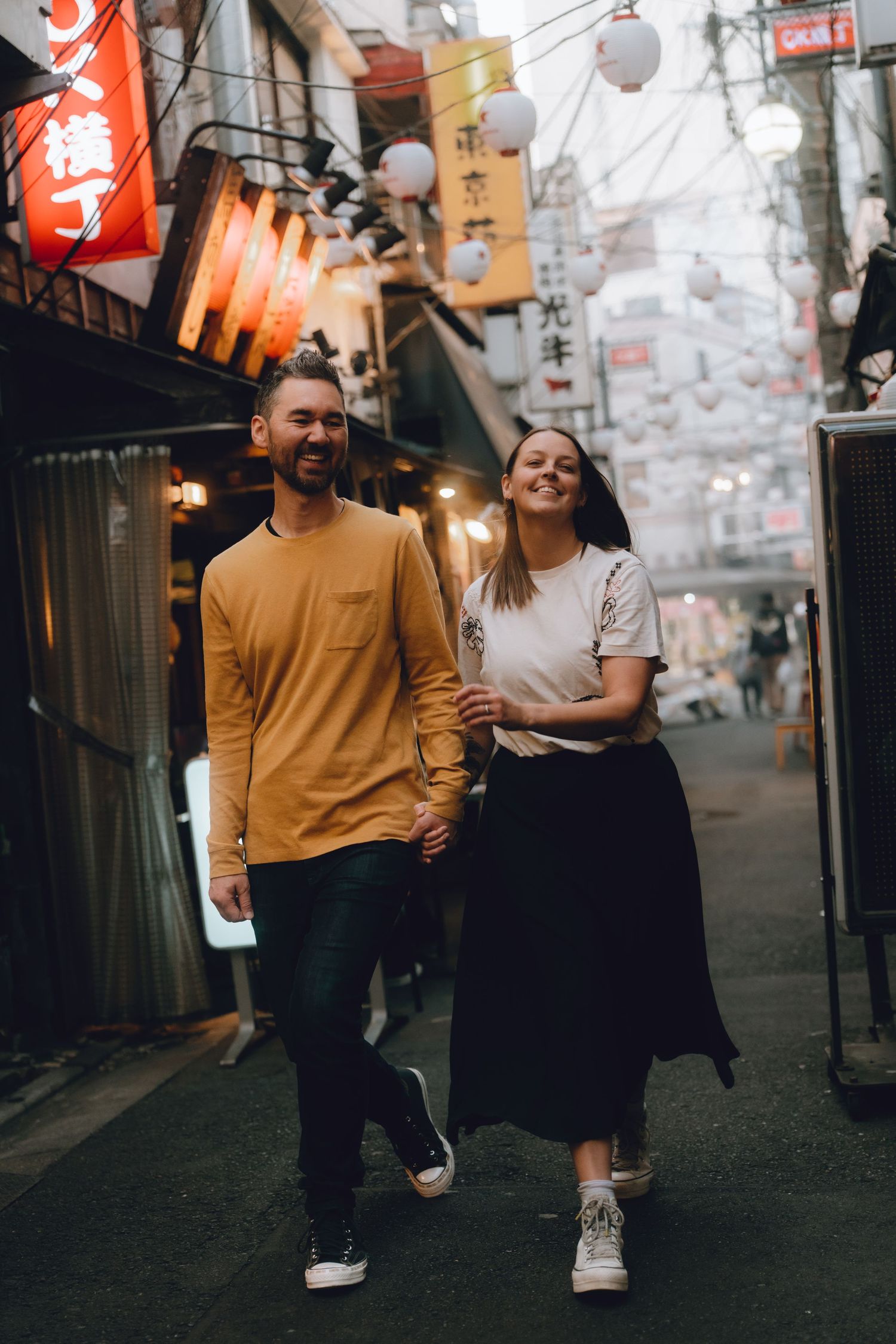 Happy couple holding hands and walking through a vibrant Japanese alley with lanterns and neon signs.