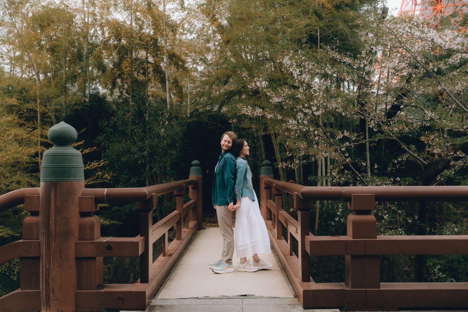Couple holding hands on a wooden bridge surrounded by lush green bamboo trees in a serene garden setting.