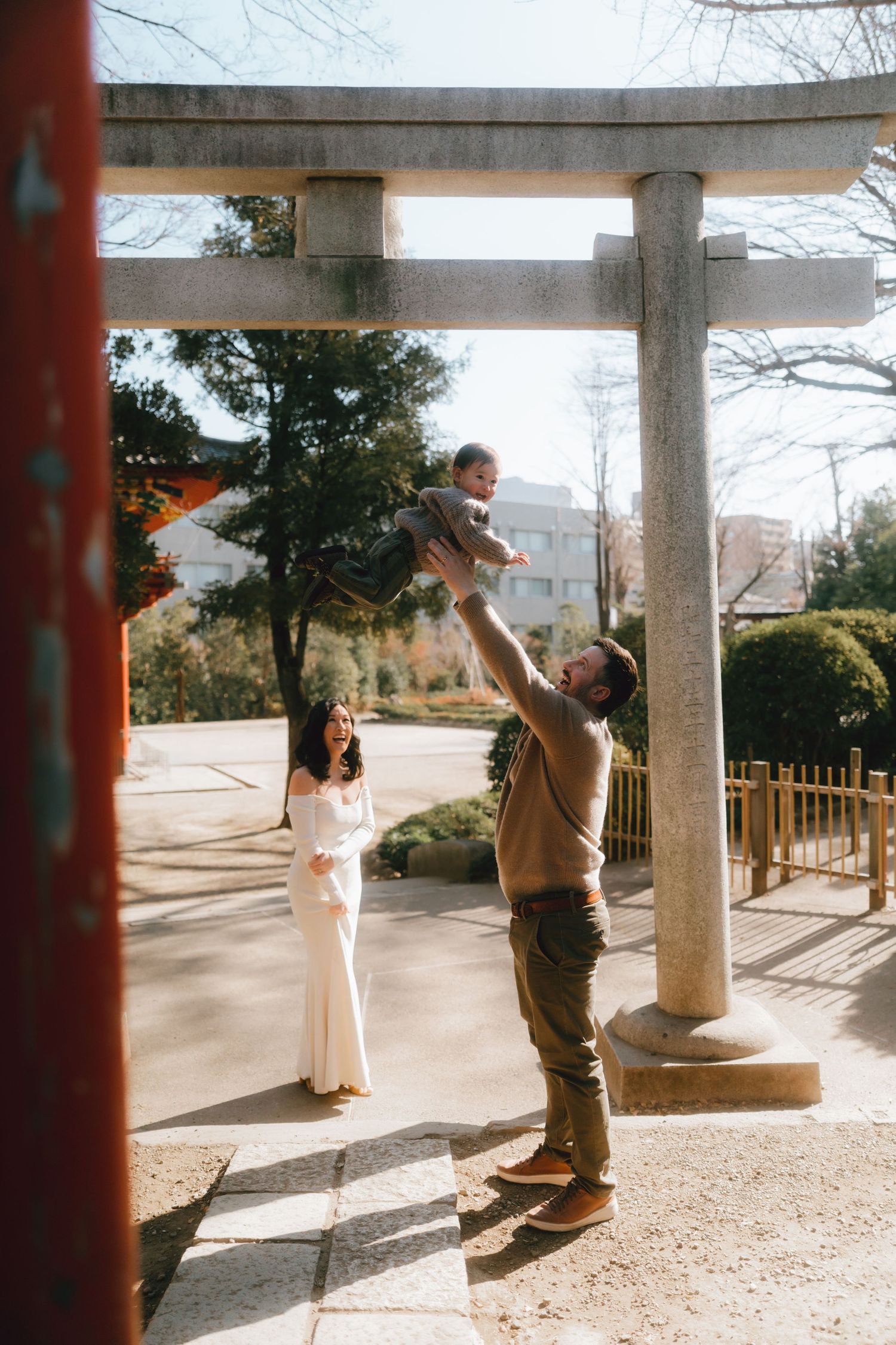 A father joyfully tosses his baby in the air near a Japanese torii gate while the mother watches in a white dress in Nezu shrine, Tokyo.