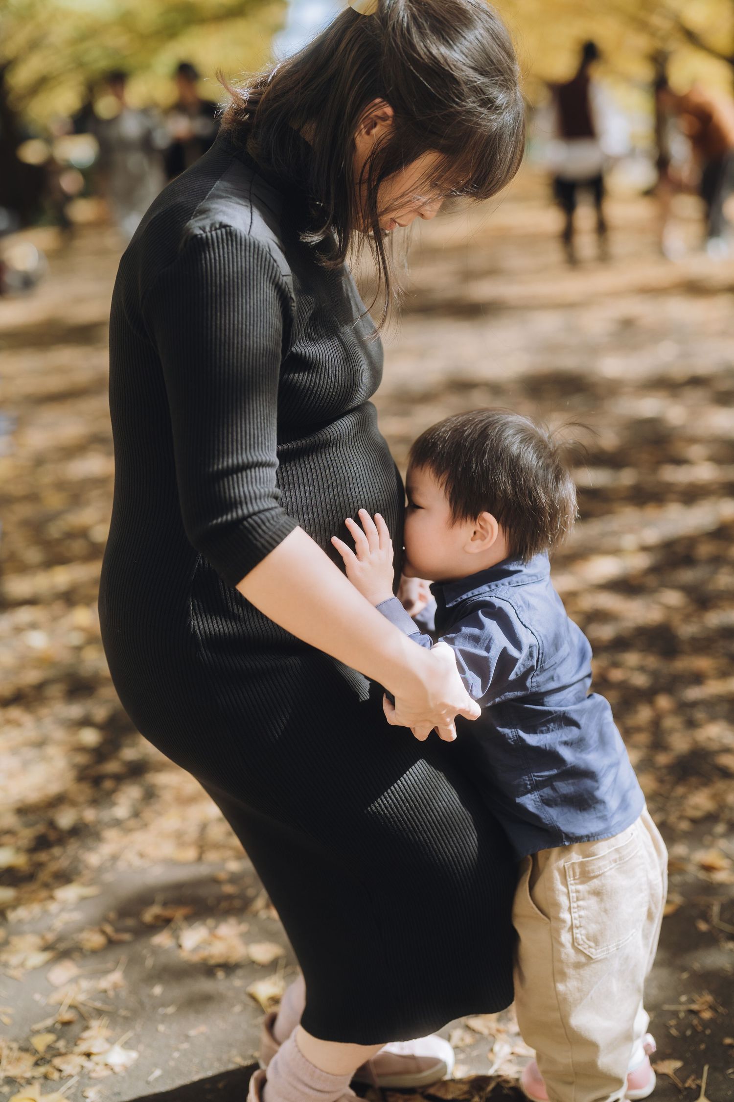 A young child hugs a pregnant woman's belly outdoors in a park during autumn.