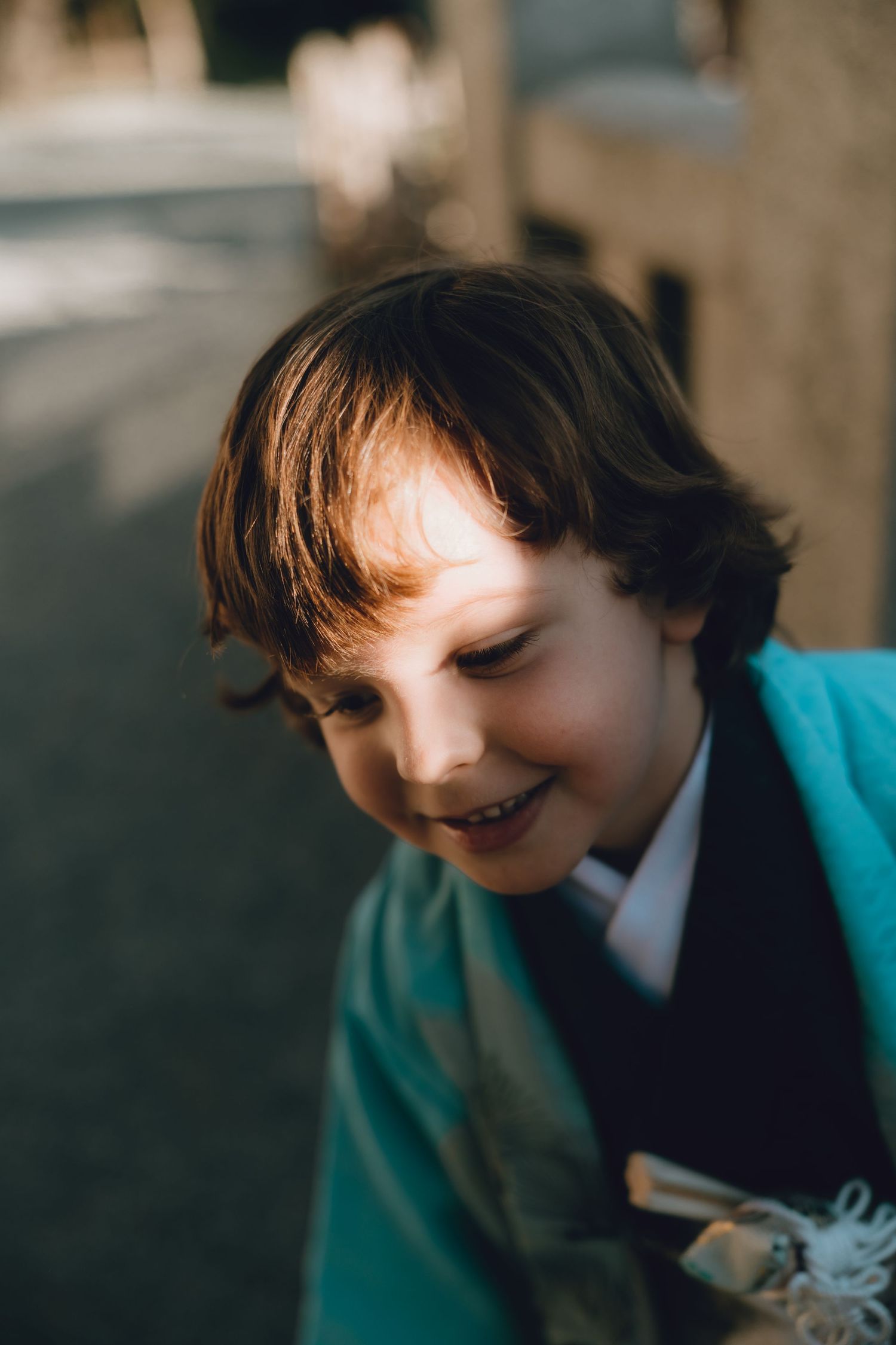A smiling young boy with brown hair wearing a teal blazer and tie, photographed outdoors in warm sunlight.