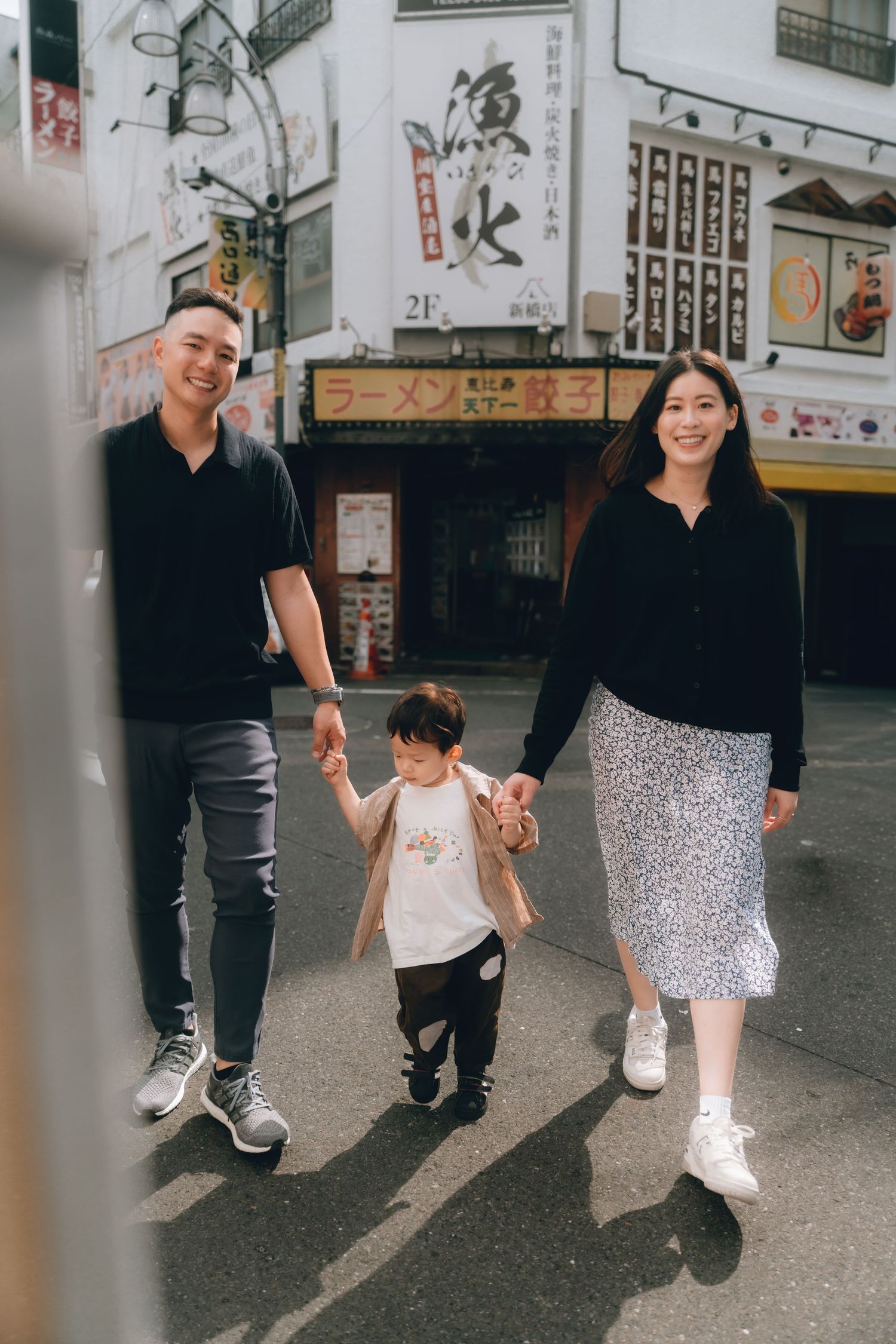A happy Asian family of three walks hand-in-hand on a Japanese street with ramen shop signs in the background in Shinbashi, Tokyo.