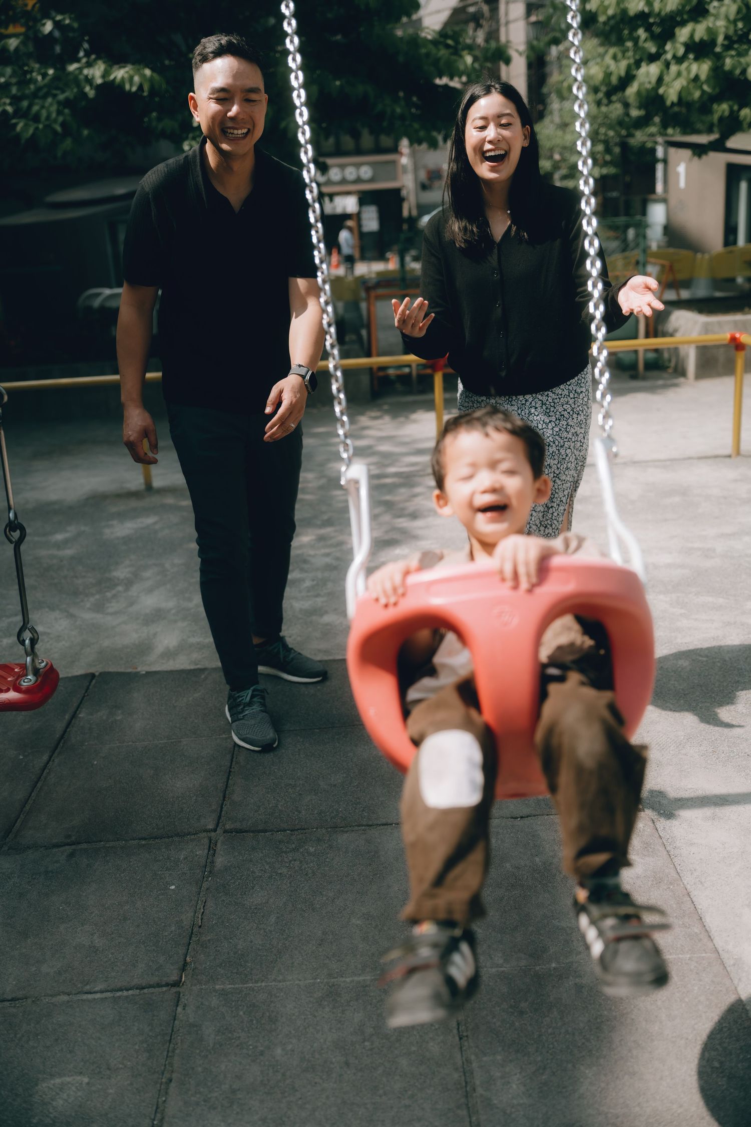 Happy Asian family at playground, parents laughing as toddler boy enjoys red bucket swing outdoors in Shinbashi, Tokyo.