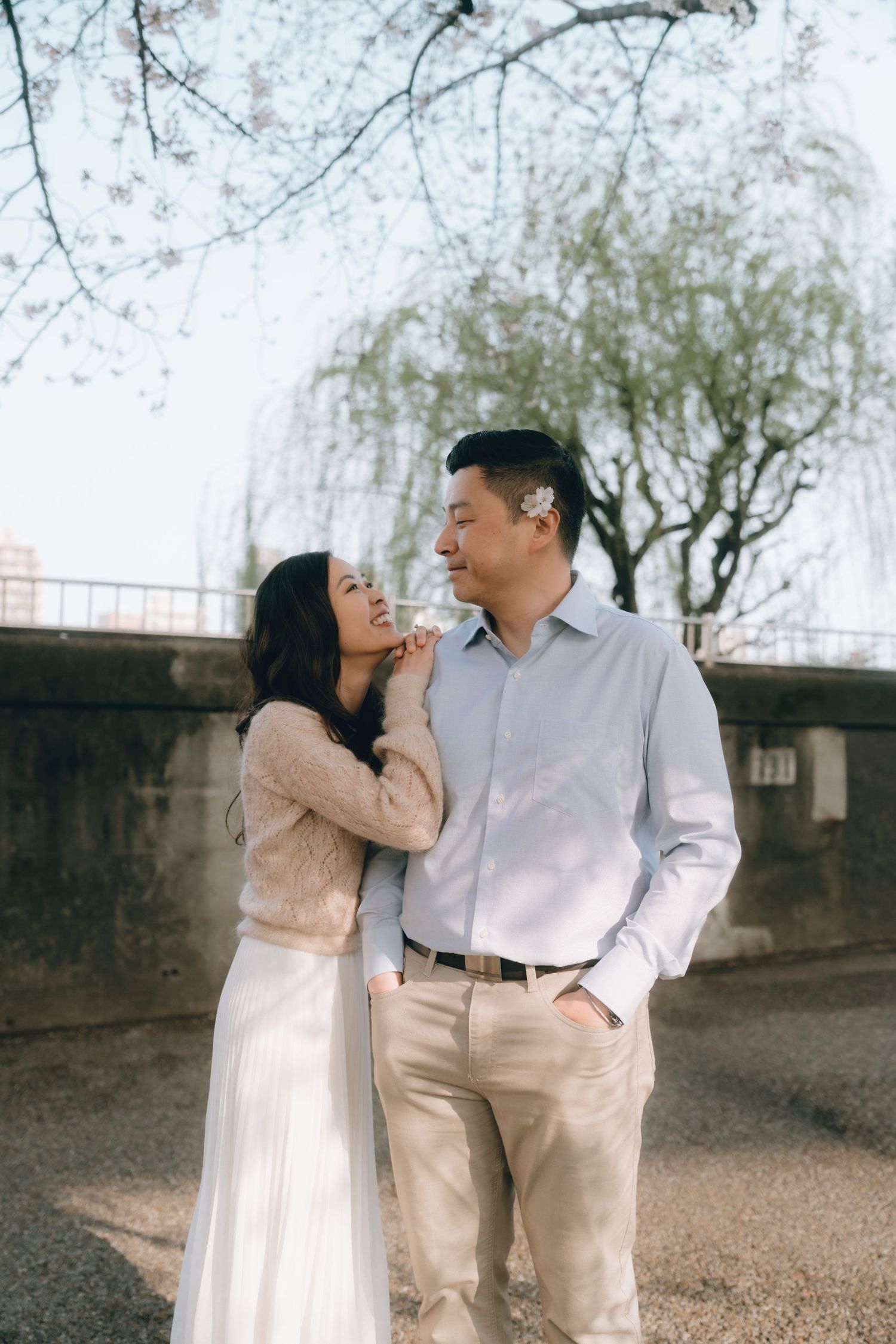 Couple sharing a romantic moment outdoors near cherry blossom trees, woman in white dress touching man's face.