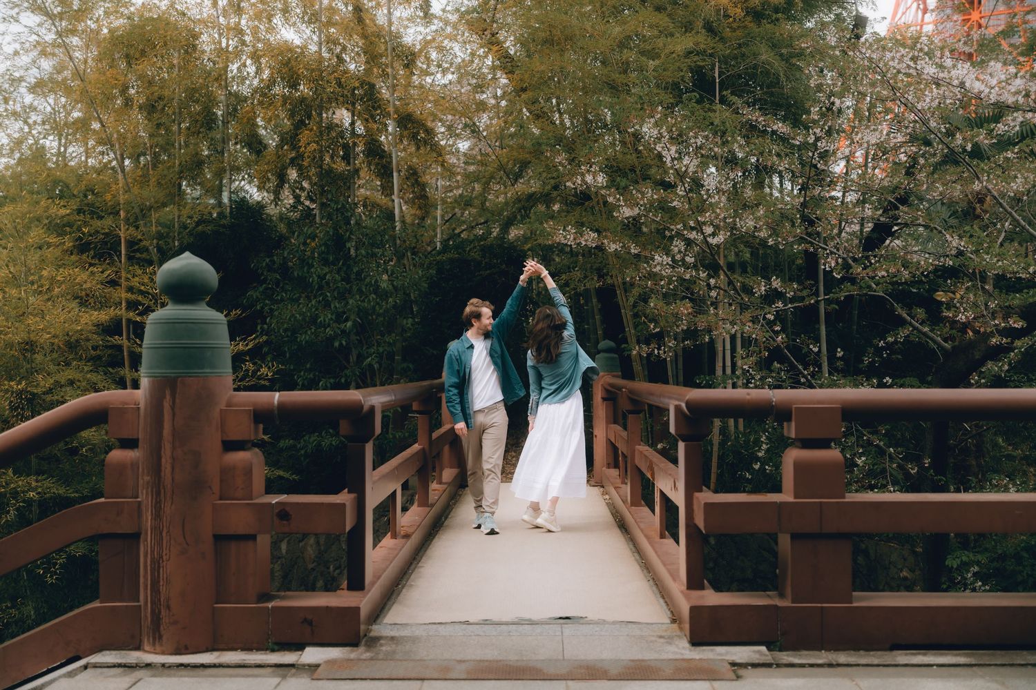 Couple holding hands walking across a wooden bridge surrounded by lush green trees in a serene garden setting.