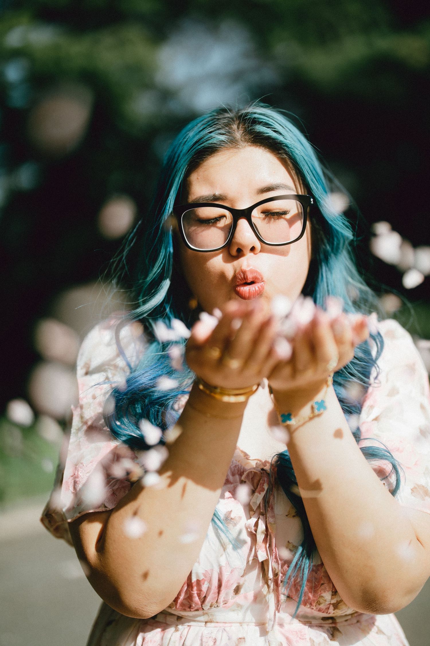 Young woman with blue hair and glasses blowing flower petals from her hands outdoors in sunlight.