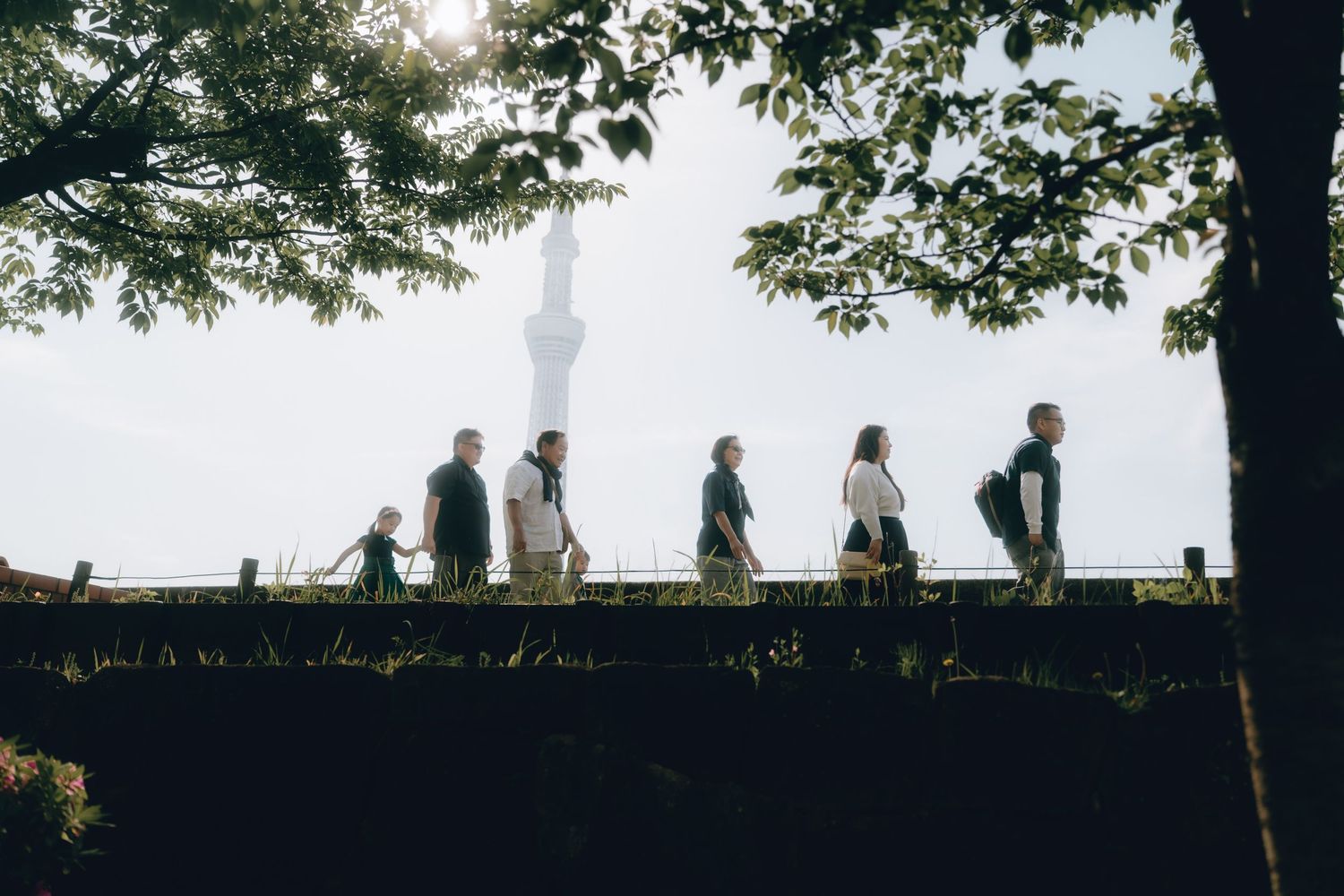 Silhouettes of people walking along a sumida park under trees with Tokyo Skytree visible in misty background.