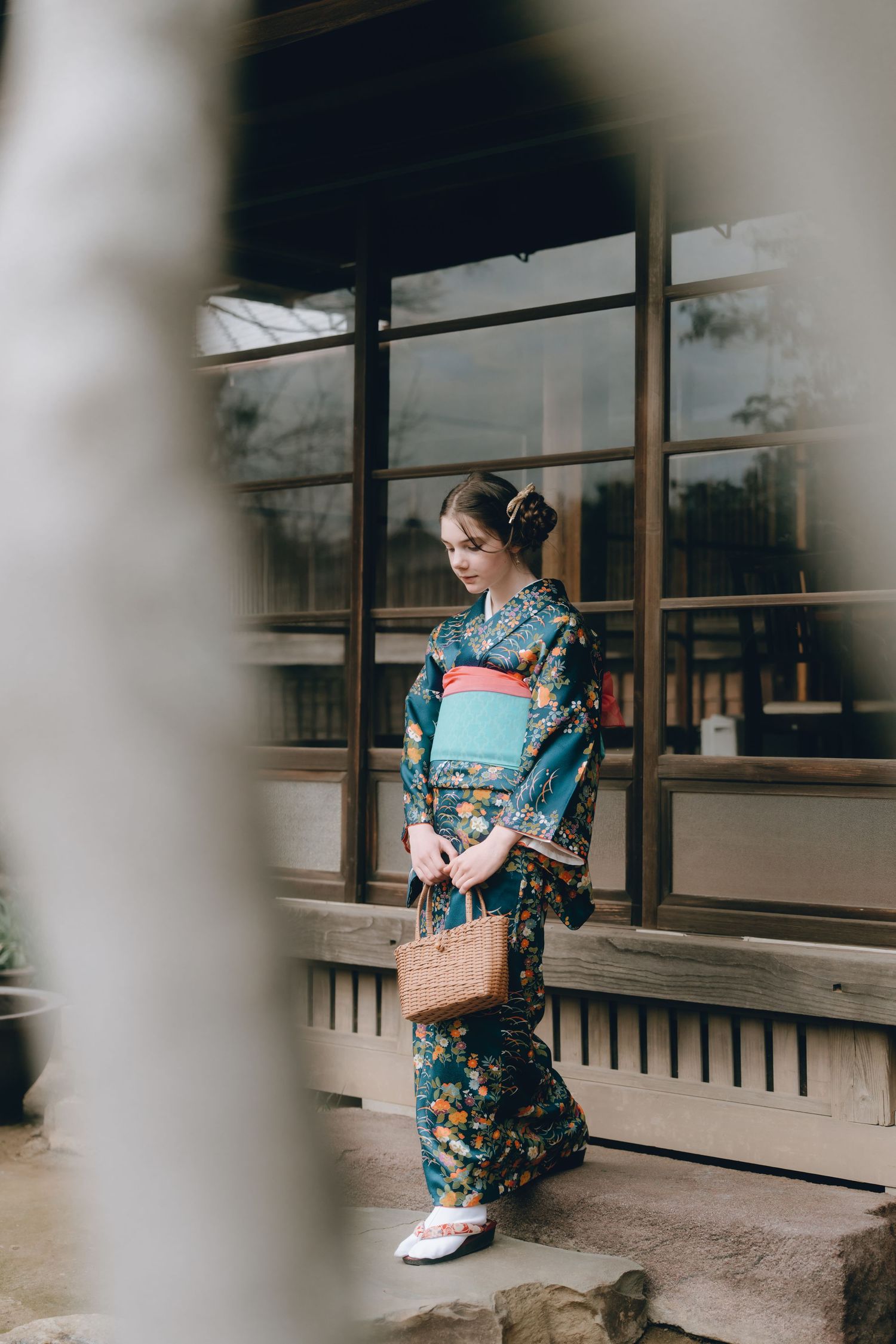 Young woman in a teal floral kimono with obi belt holds a wicker bag outside a traditional Japanese building in Sawara, Chiba, Japan.