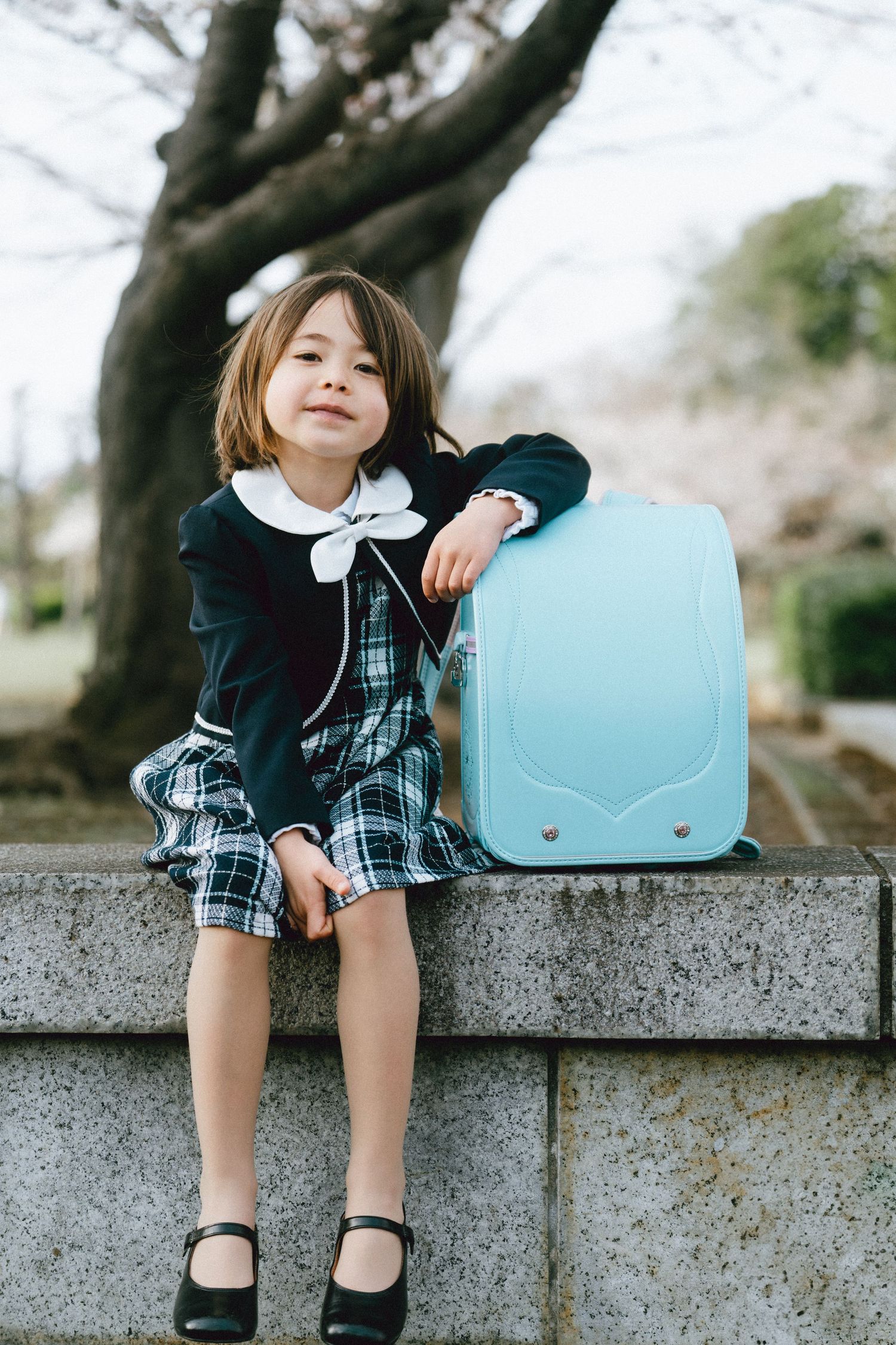 Young girl in school uniform sitting on stone wall with light blue randoseru backpack outdoors.