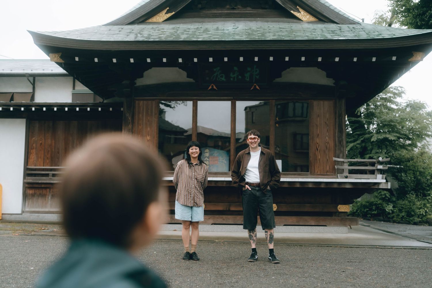 Two people stand smiling in front of a traditional Japanese shrine while a child stands in the foreground in Shimokitazawa, TOKYO.