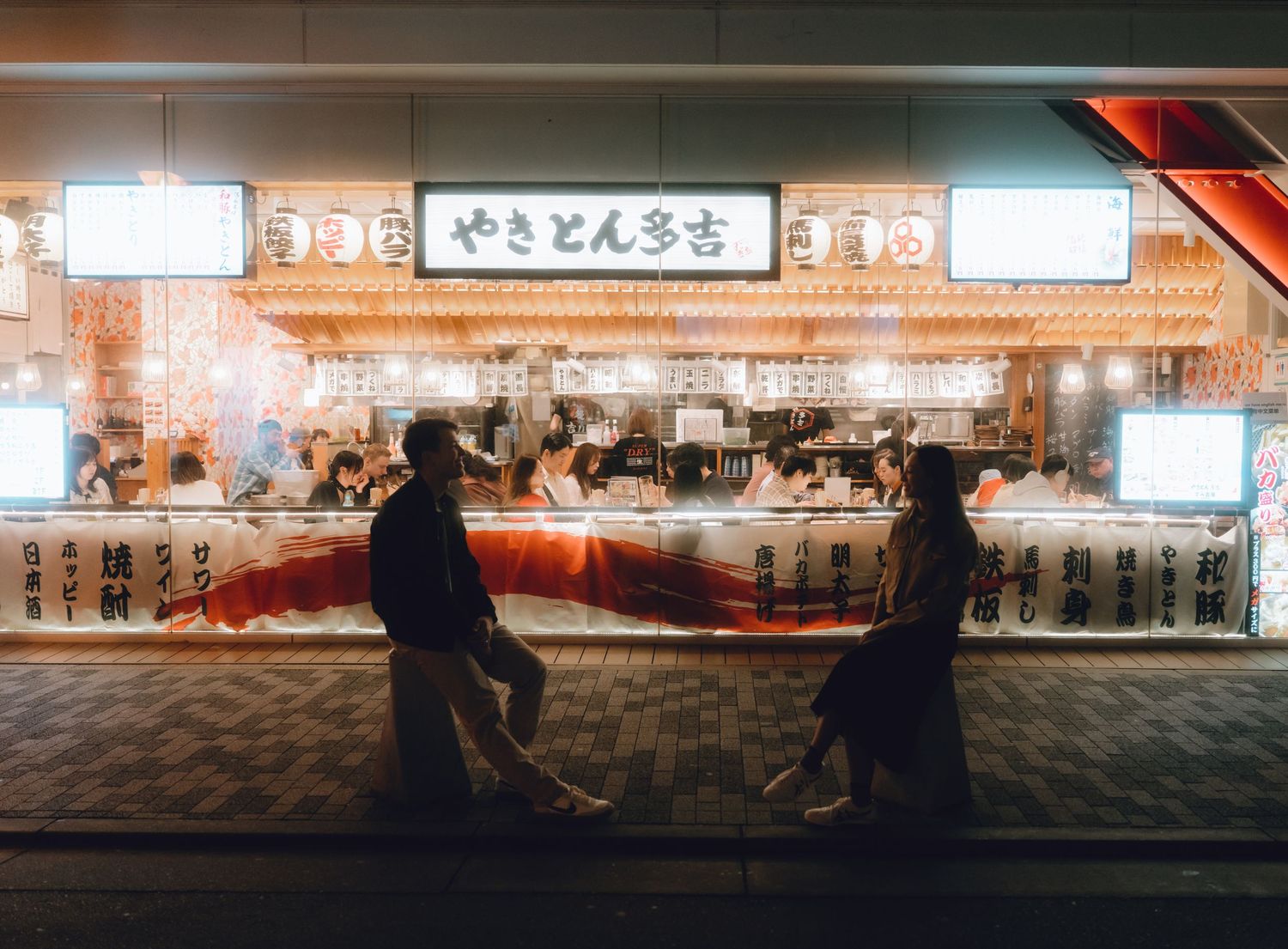Two people carry a large red fish outside a glowing Japanese yakitori restaurant at night in yurakucho.