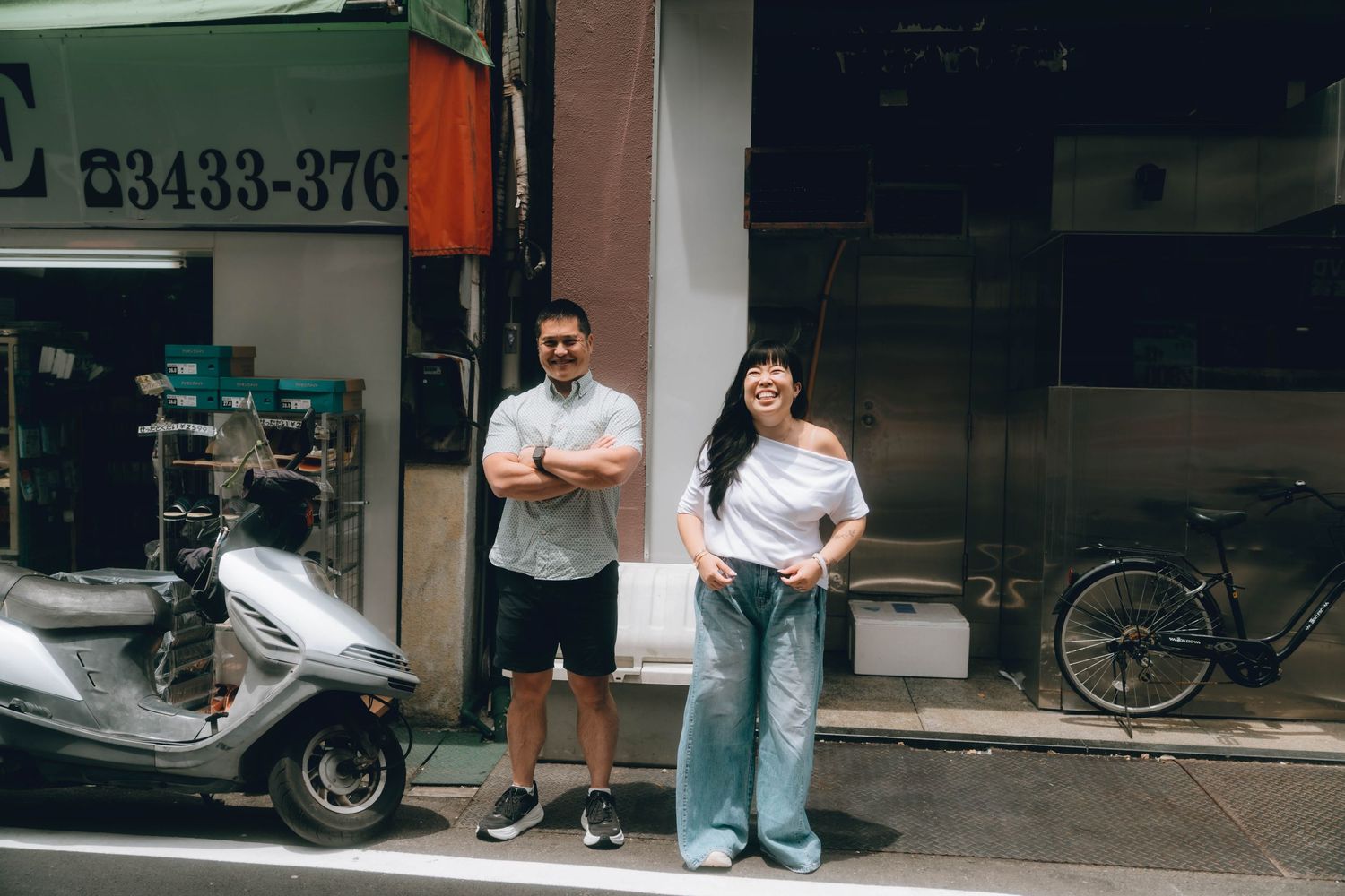 A man and woman stand outside a urban storefront in casual clothing, with a scooter and bicycle parked nearby.