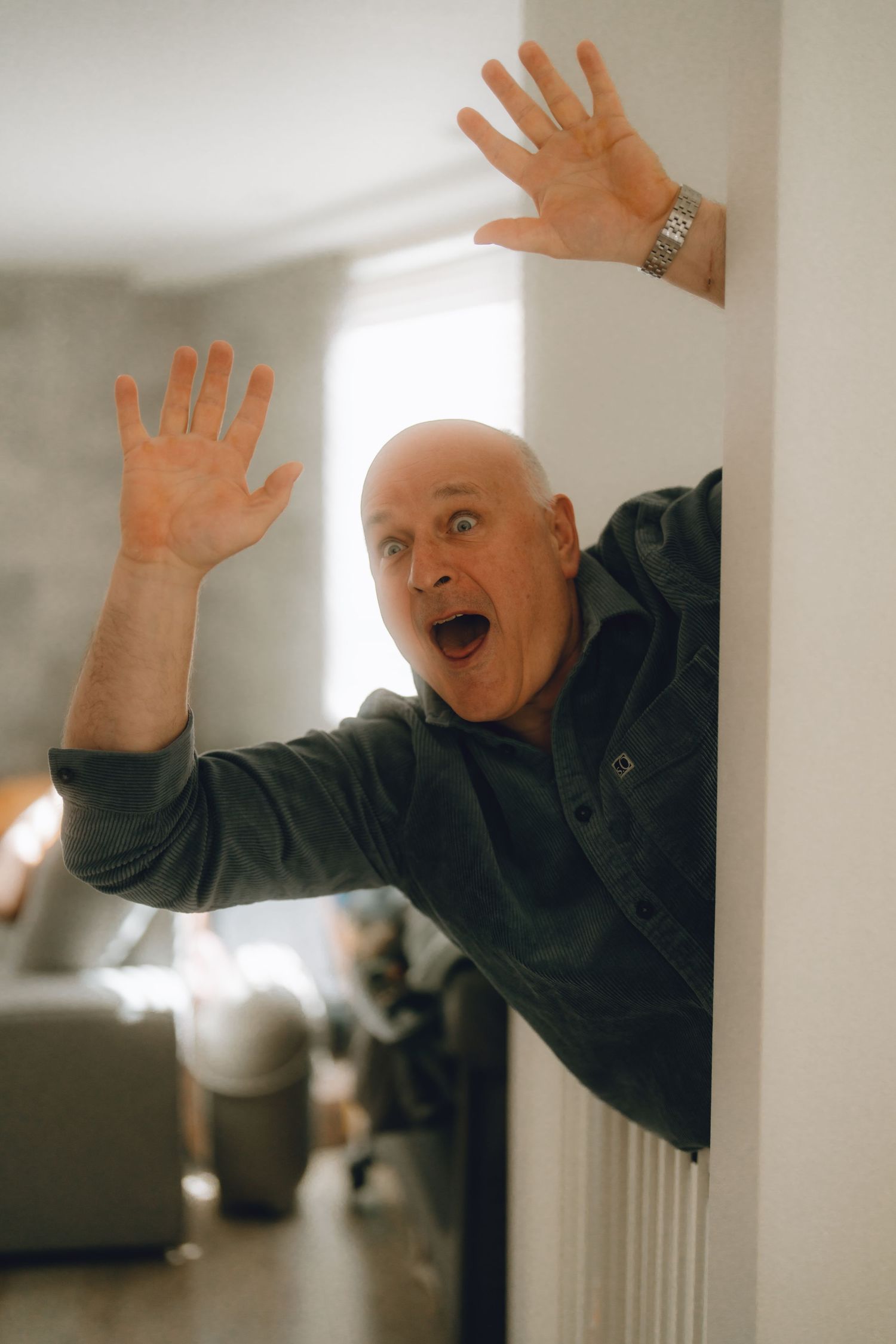 Bald man peeking around a white wall with hands raised and mouth open in a surprised expression indoors.