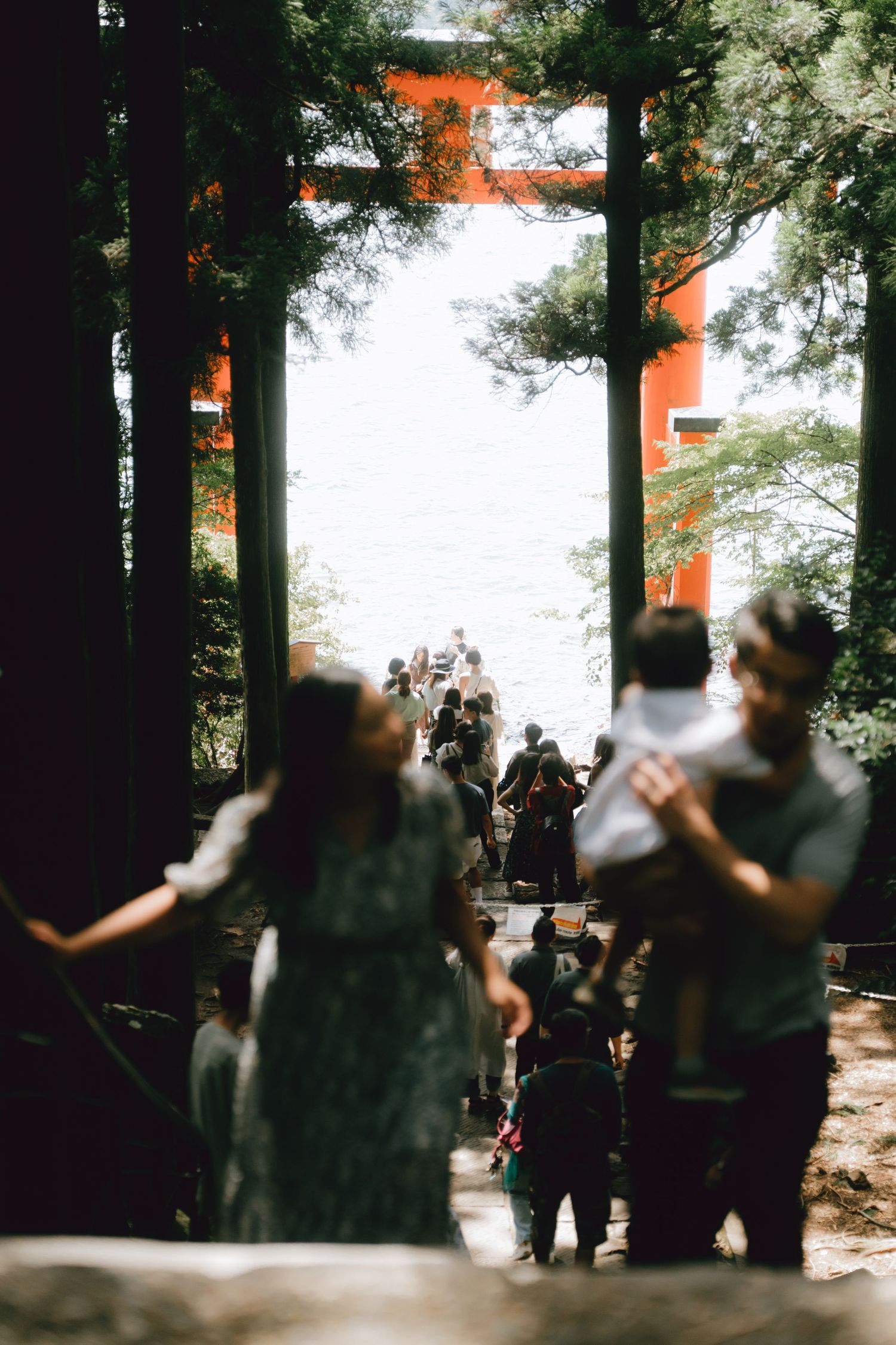 Visitors walking through a forested Japanese shrine path toward a red torii gate near water.