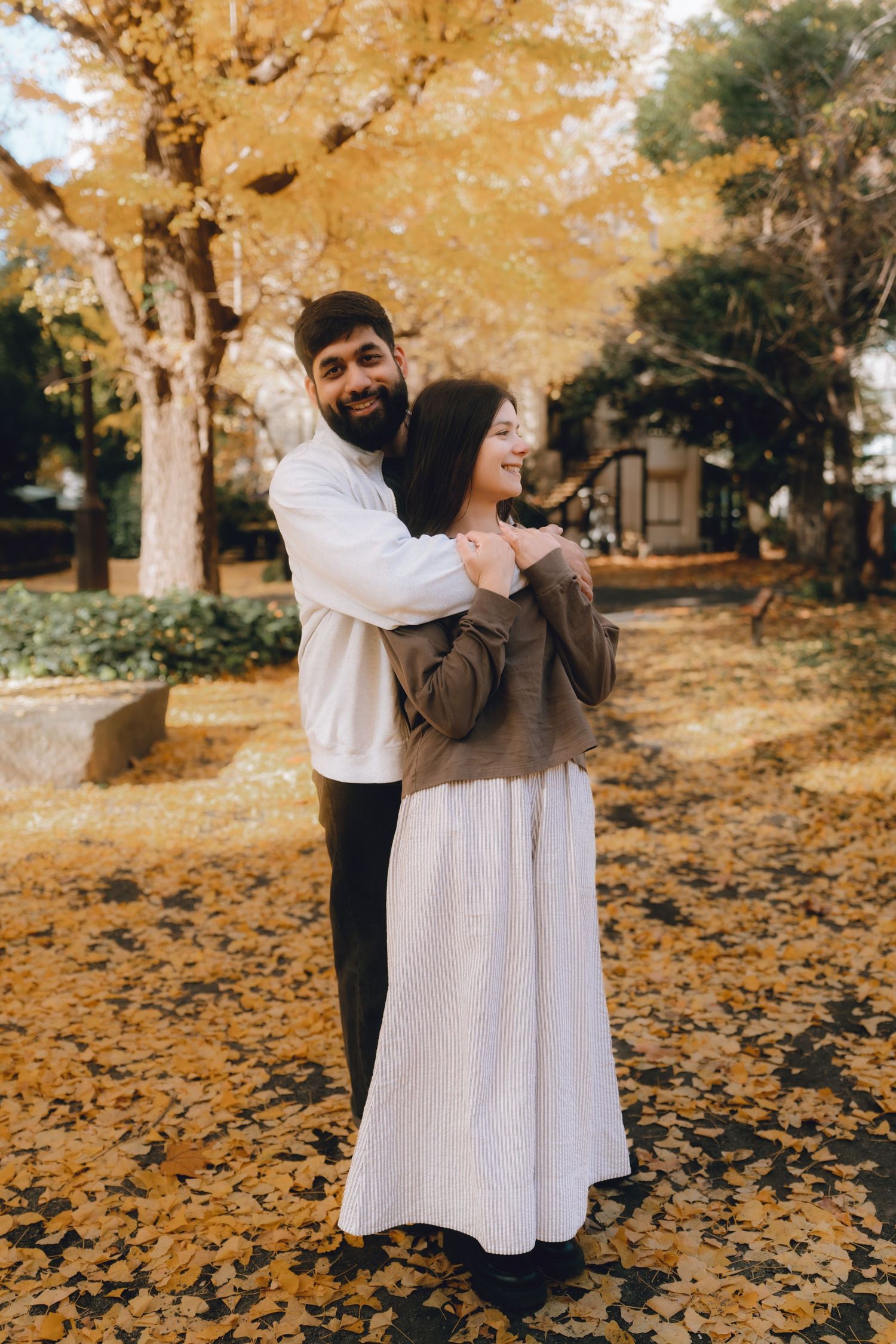 Couple embracing in autumn park with golden ginkgo leaves covering ground and trees in warm fall colors.