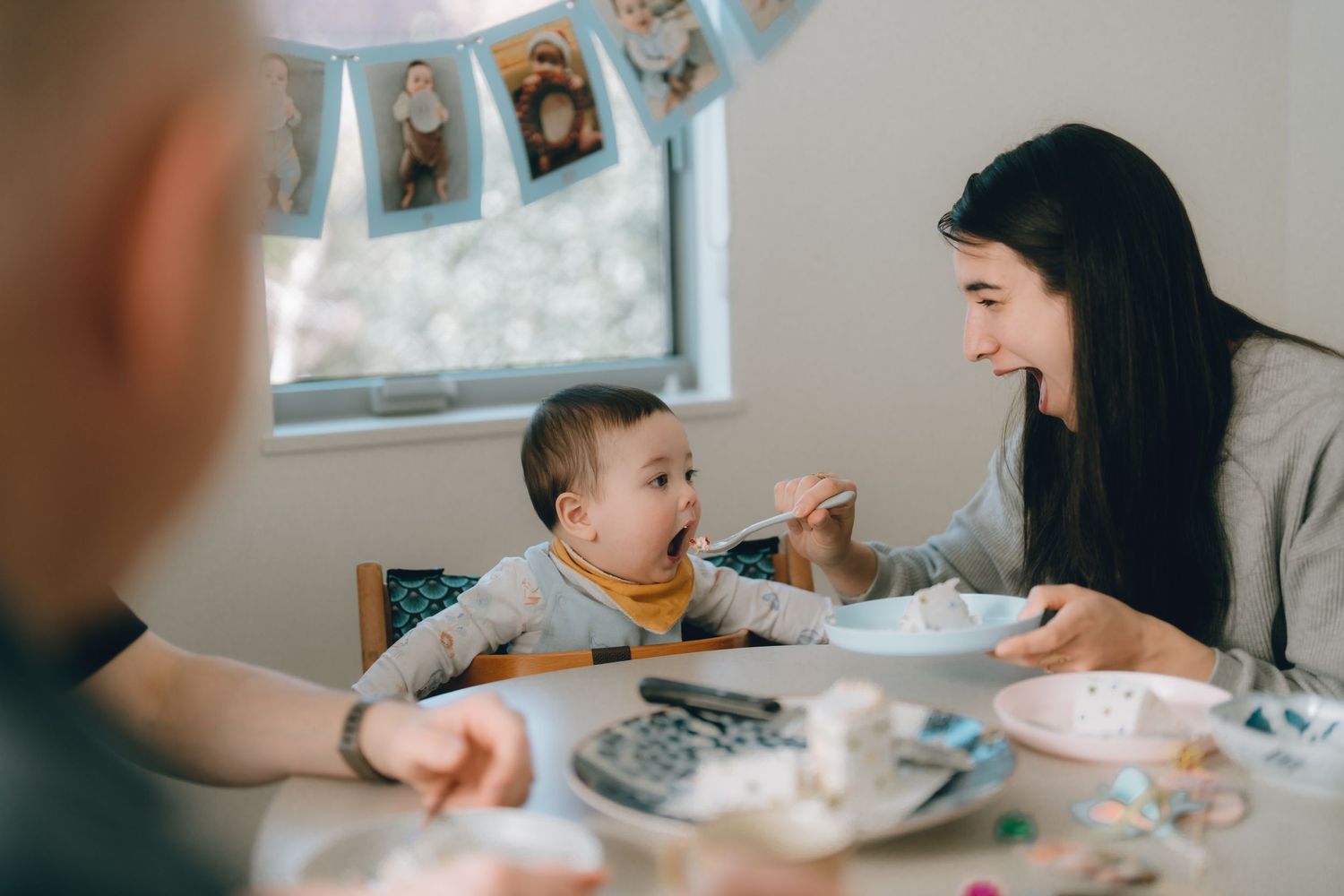 A smiling mother feeds her baby in a high chair at a family dining table decorated for a birthday celebration.