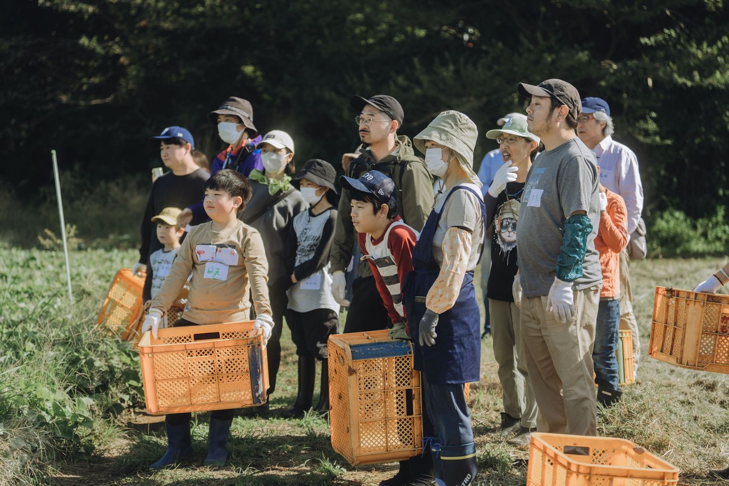 Group of adults and children holding orange crates during an outdoor farm harvest activity in Japan.
