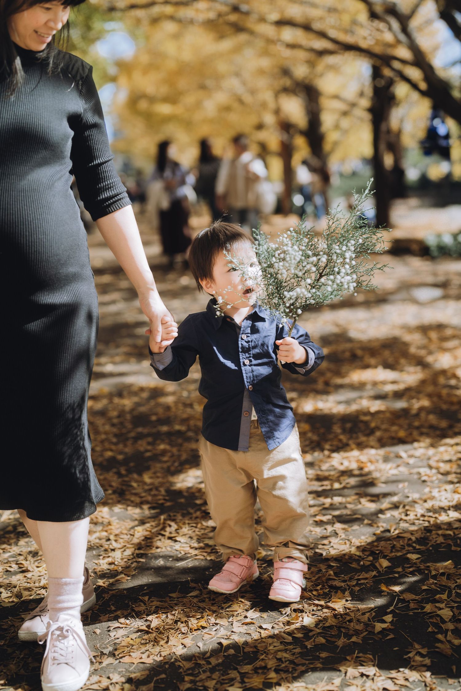 A toddler in a denim jacket holds a small plant while walking hand-in-hand with a woman in an autumn park in Tokyo.