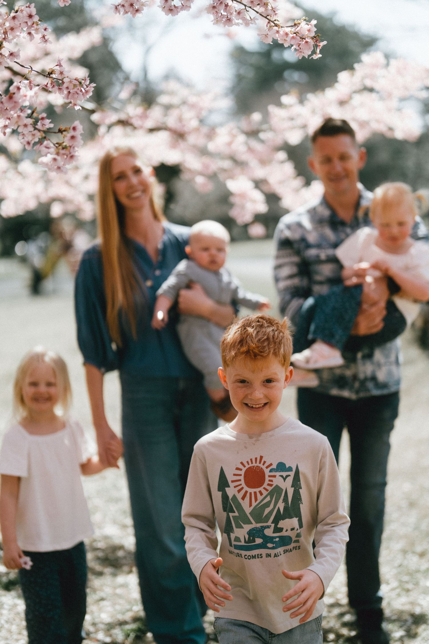 Family of five posing under cherry blossom tree in spring, with redhead boy smiling in foreground.