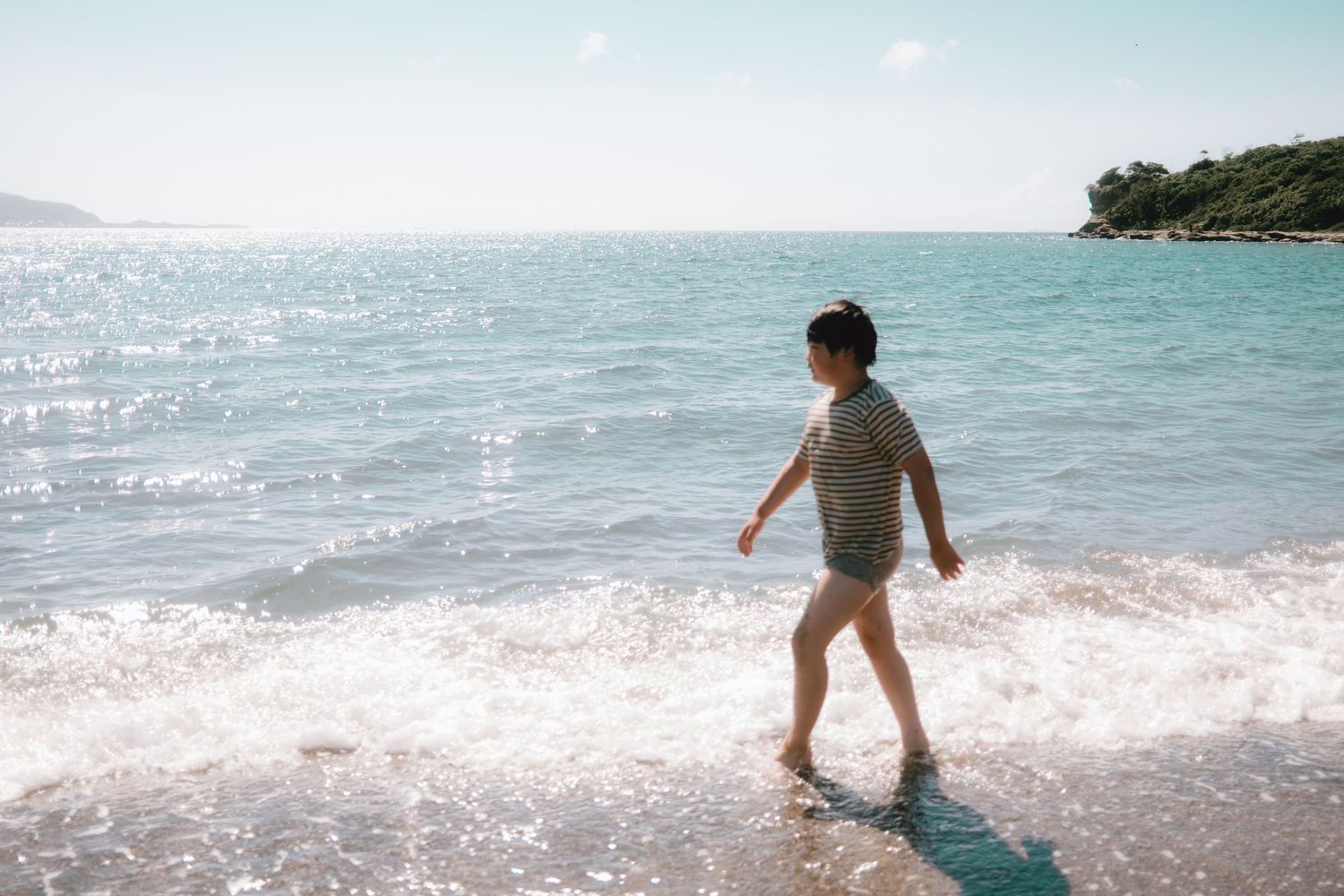 A young child walks along a sunny beach shoreline with gentle waves and turquoise sea water in the background. Tateyama, Chiba.