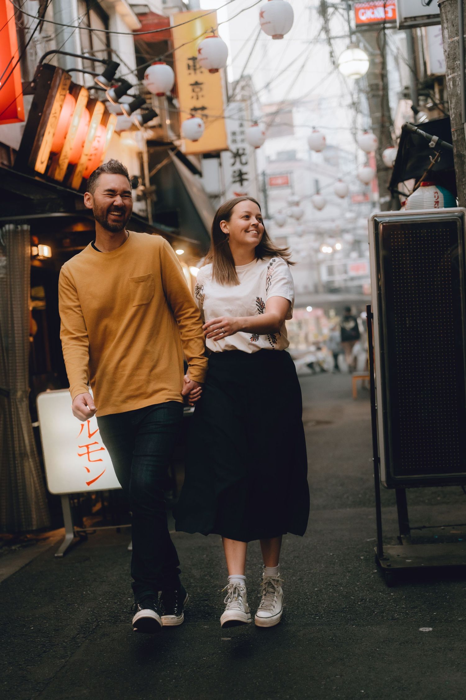 Smiling couple walking hand in hand through a vibrant Japanese alley with lanterns in tokyo.