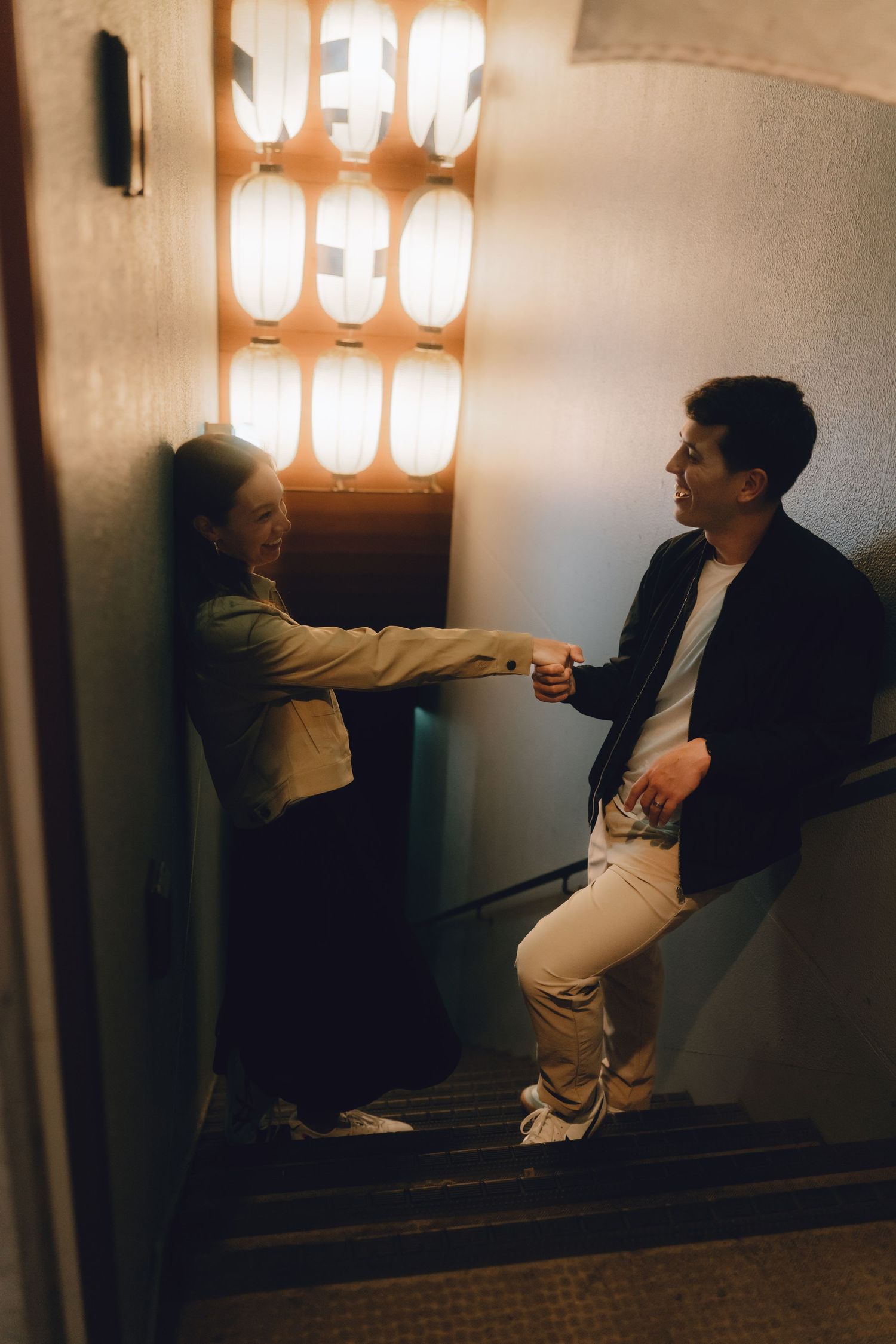 Couple holding hands on dimly lit staircase with glowing Japanese paper lanterns in background.