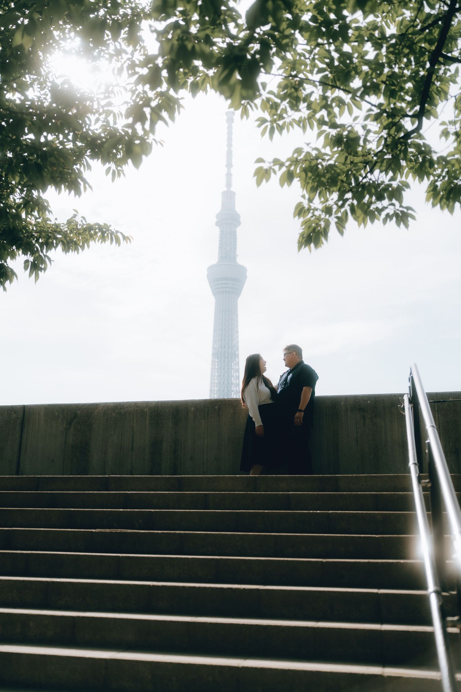 Couple standing on steps with Tokyo Skytree tower rising behind them, framed by green tree branches.