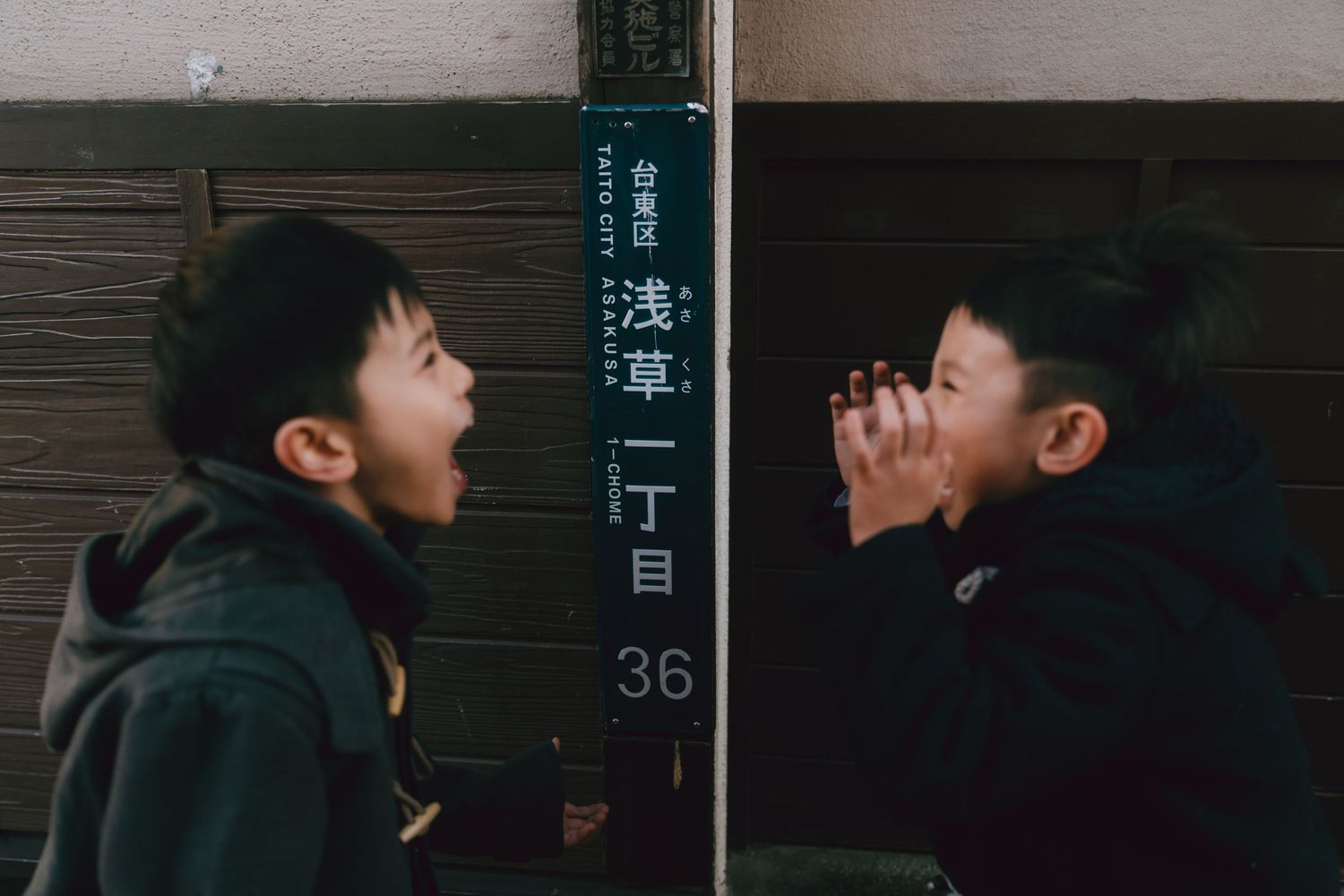 Two young boys facing each other with open mouths near a Japanese street sign in Asakusa, Tokyo.