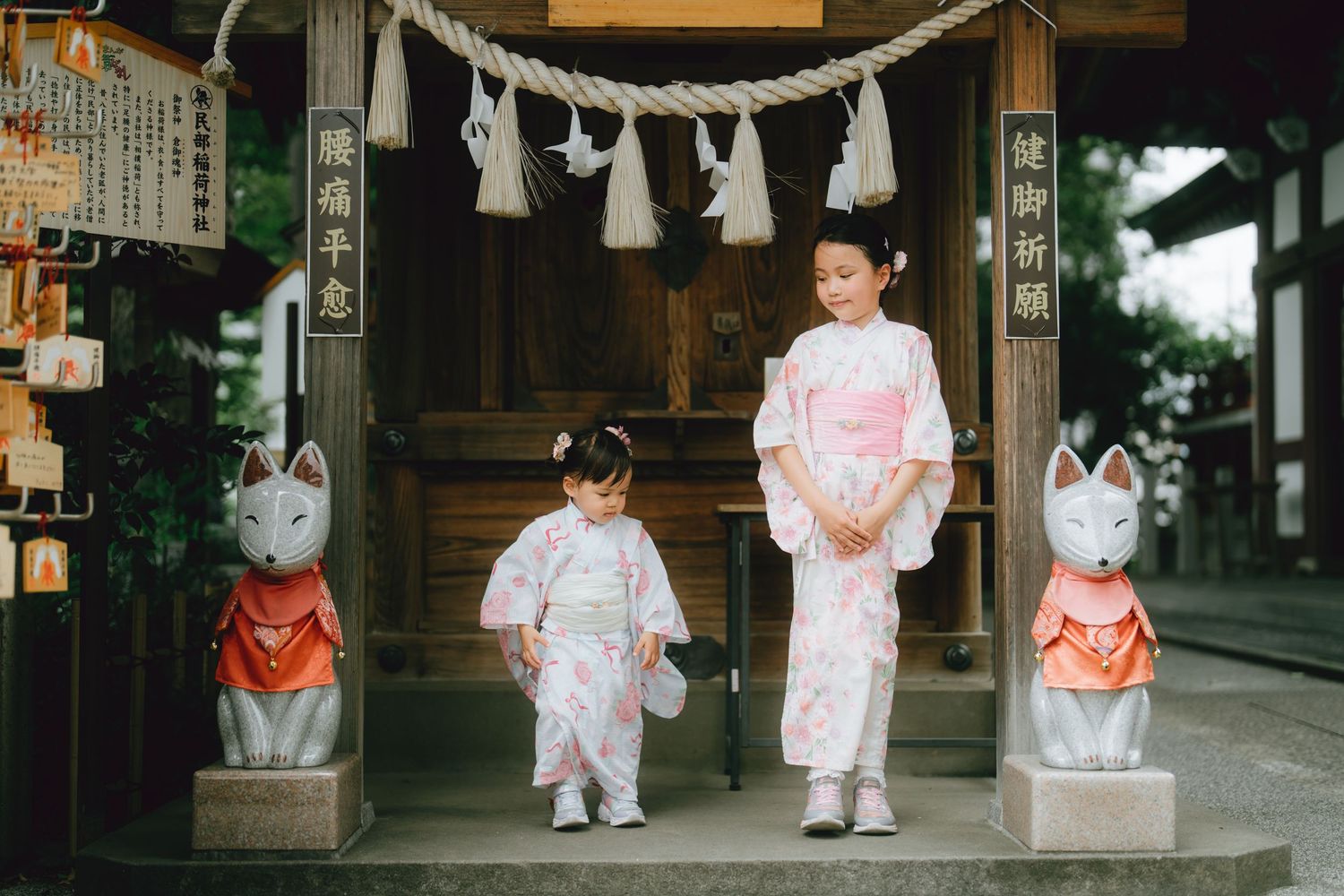 Two girls in pink floral kimonos pose at a Japanese shrine in Yokohama flanked by fox statues.