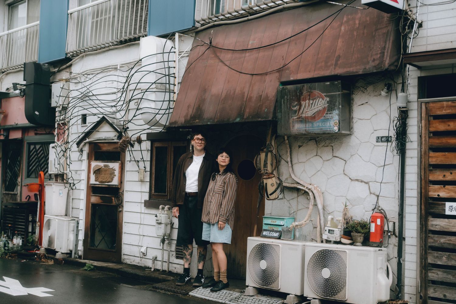 Couple standing in front of a vintage weathered building with tangled wires and an air conditioning unit on a rainy day shimokitazawa,tokyo,