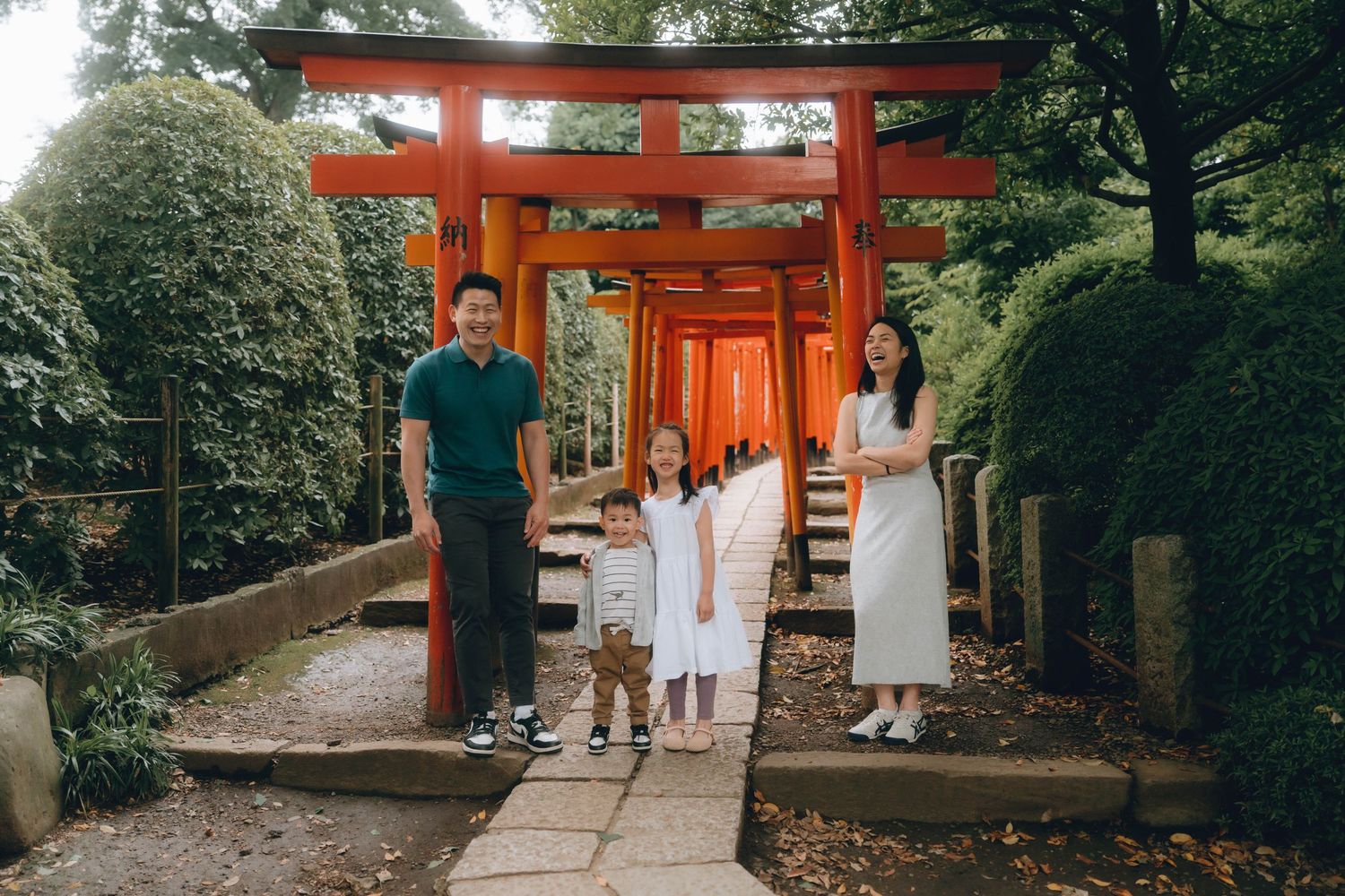 Family of four posing under red torii gates in in Nezu shrine, Tokyo. , with lush greenery and stone path.