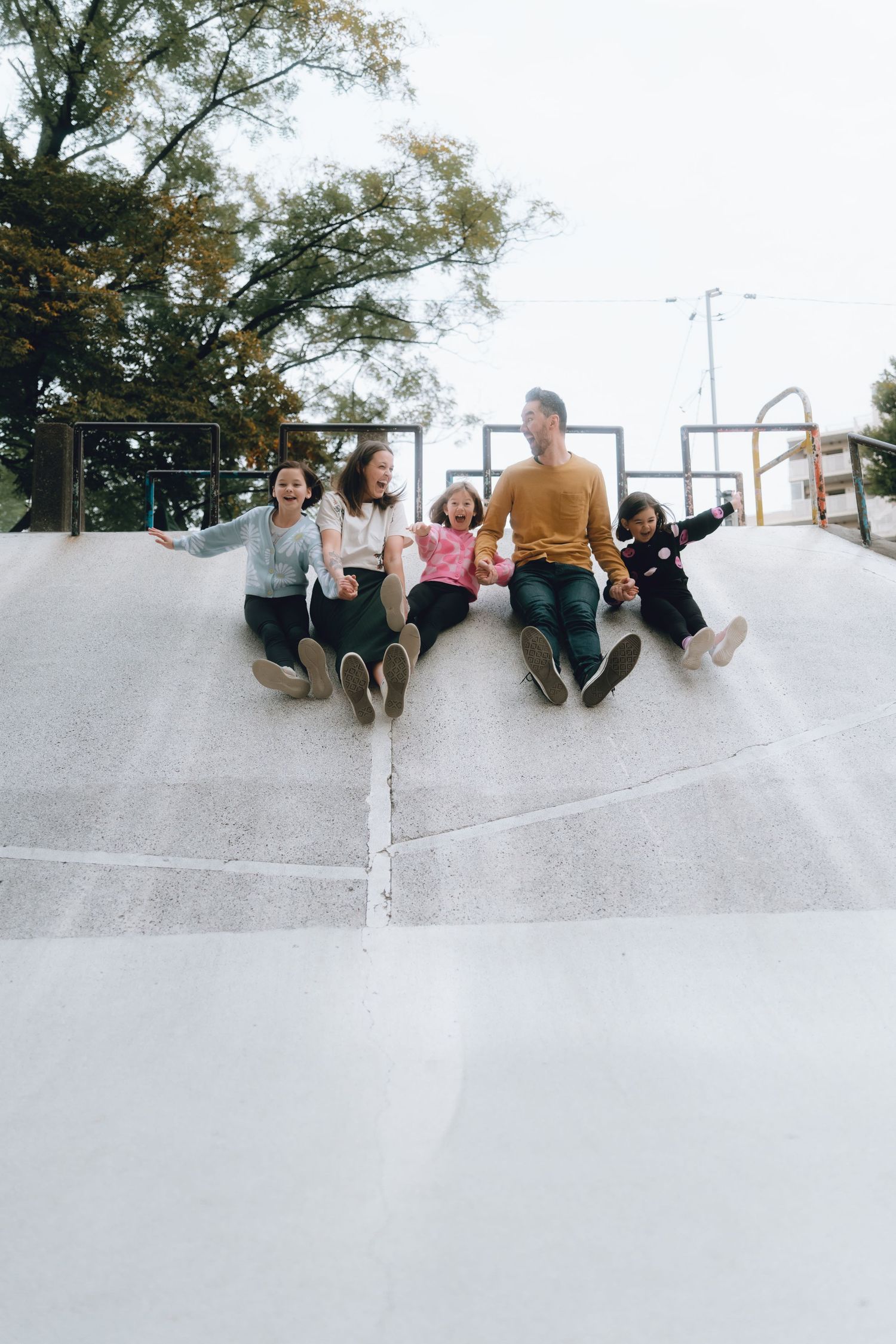 Happy family of four sitting and sliding down a concrete skate park ramp outdoors, smiling with arms raised.
