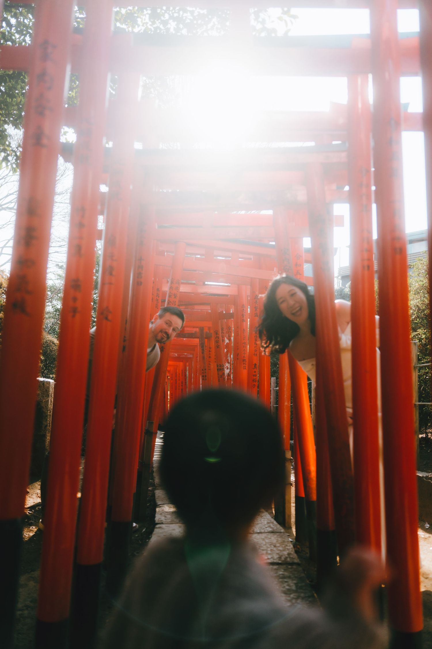 Couple posing playfully among vibrant red torii gates at a Japanese shrine, with sunlight streaming through in Nezu shrine.